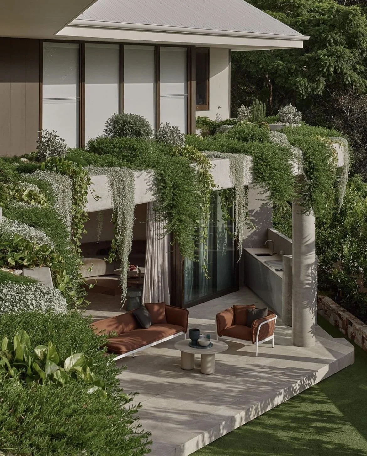 Modern house with a balcony filled with lush green plants, overlooking a patio with outdoor furniture and a garden.