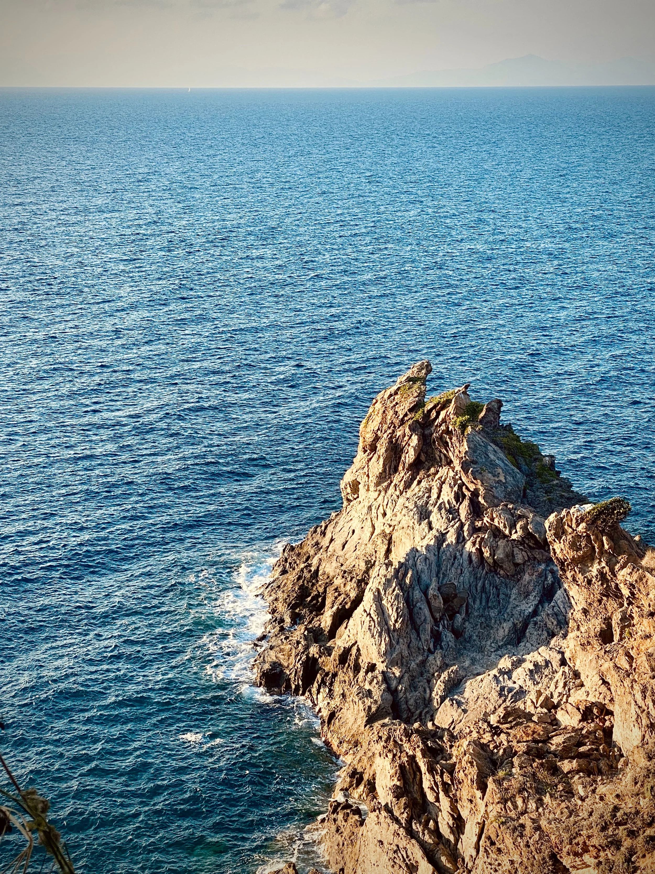 A rocky coastline with a large, jagged rock formation extending into the ocean, with the water appearing calm and blue, and a distant horizon with faint landmasses.