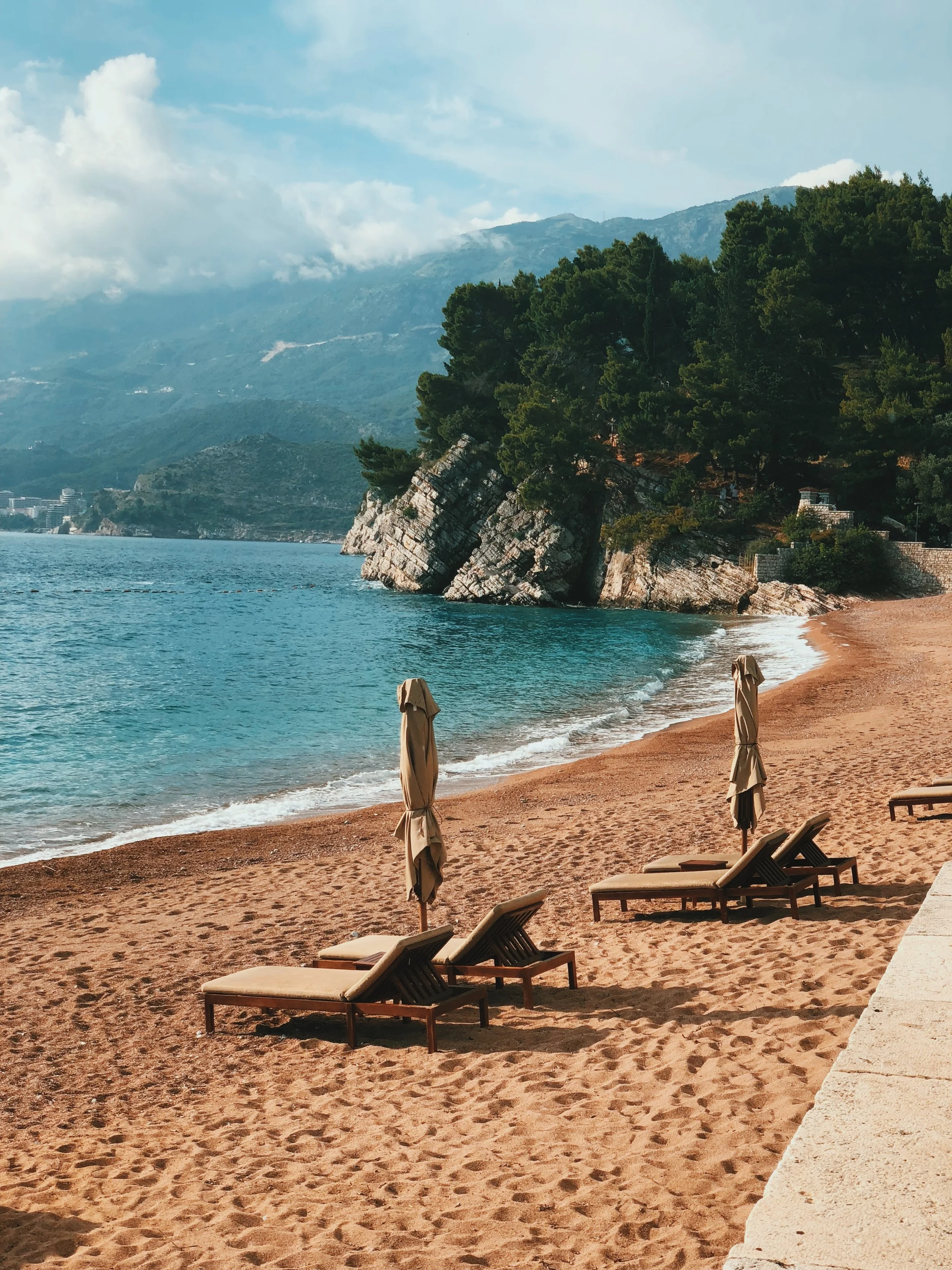 Empty beach with two loungers and umbrellas, rocky cliffs, forested hills, and mountains in the background under partly cloudy sky.