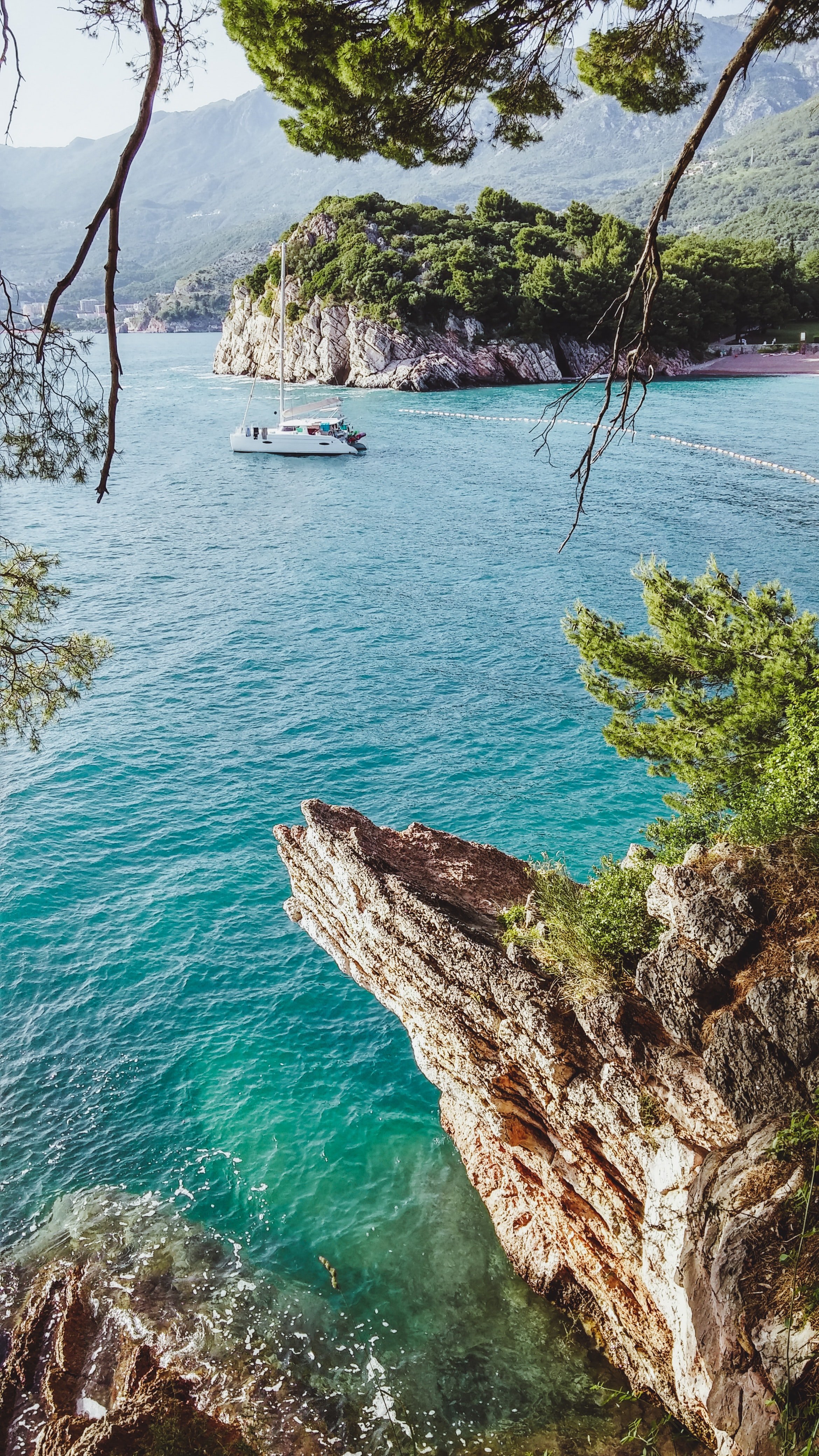 A scenic view of a turquoise sea with a white sailboat near a rocky, green-covered island. The photo is framed by trees with green foliage and a rocky shoreline.