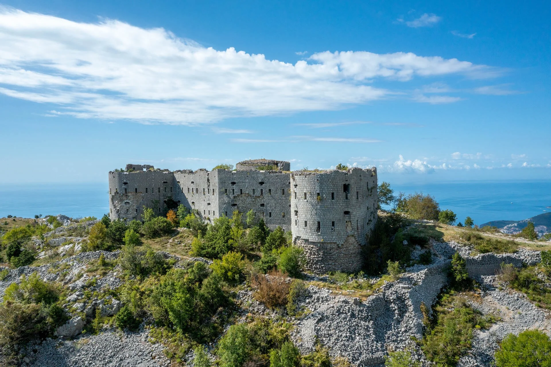 Ancient stone castle on a hilltop surrounded by greenery with a view of the ocean and blue sky in the background.