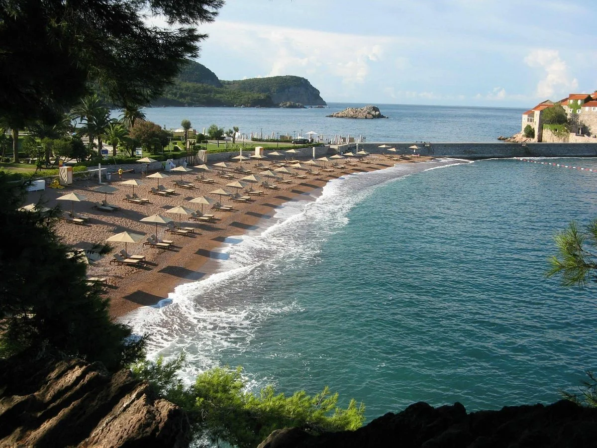 Beach with rows of empty lounge chairs and umbrellas, calm sea, rocky outcrop in distance, trees at the shoreline