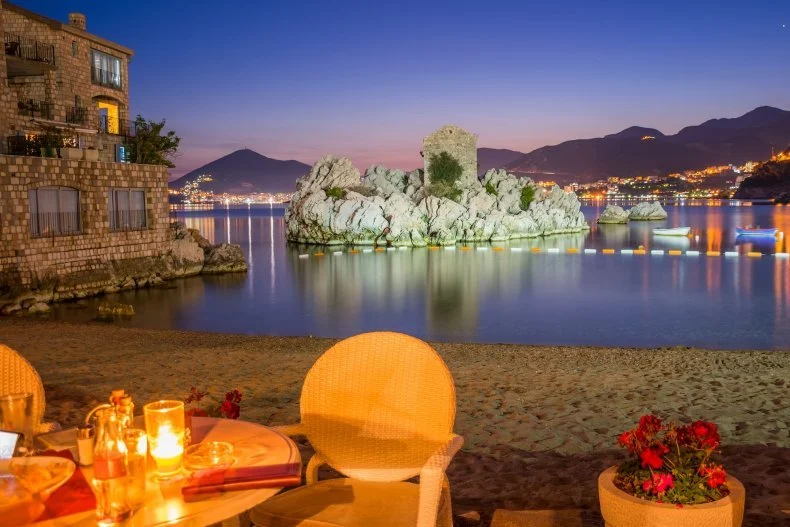 Beachside restaurant at dusk with a view of rocks in the water, buildings on a hillside, and mountains in the background.