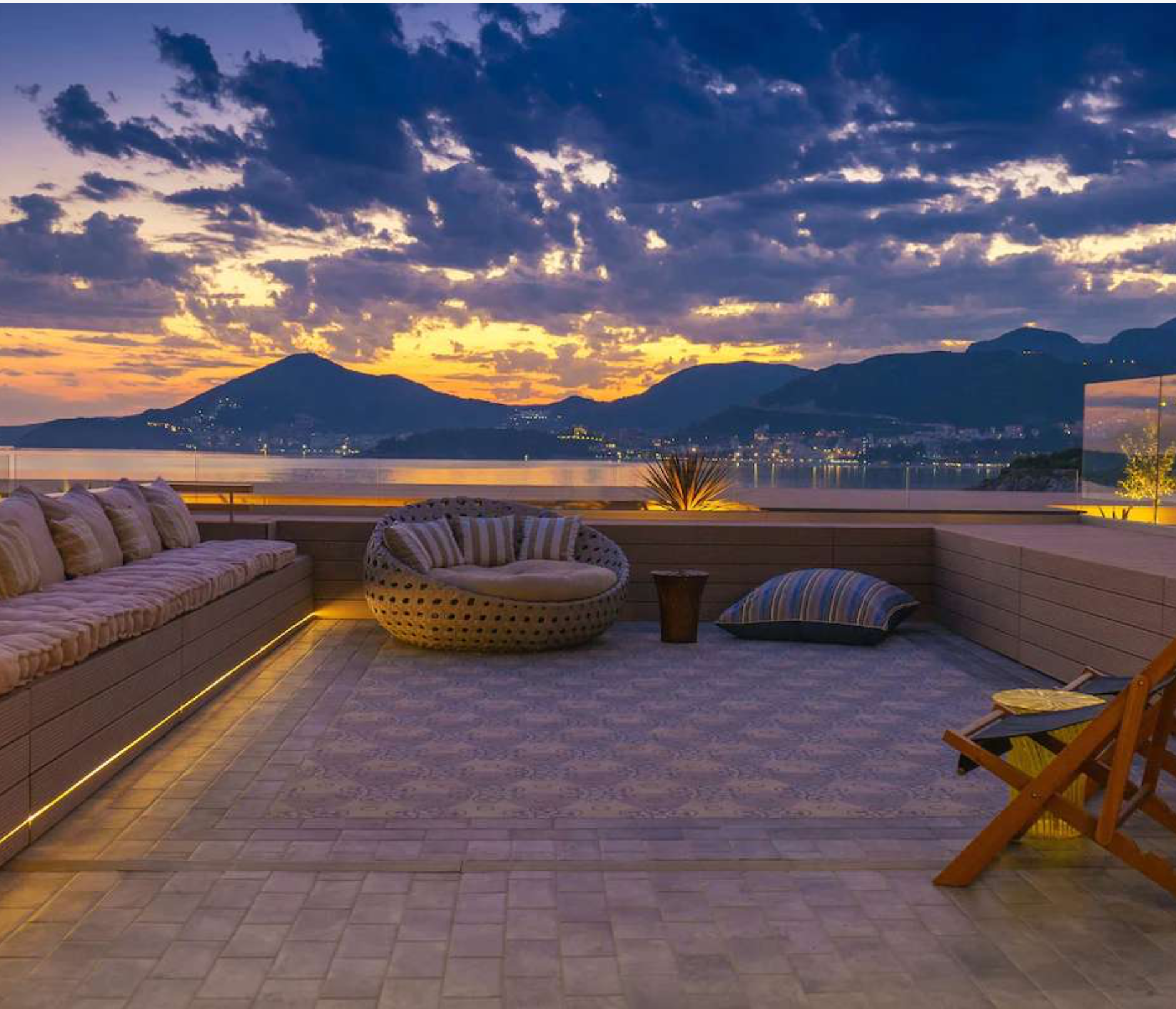 A rooftop patio with outdoor furniture overlooking a lake and mountain range during sunset, featuring a long cushioned bench, a round woven chair, a small table, striped pillows, and a wooden deck chair.