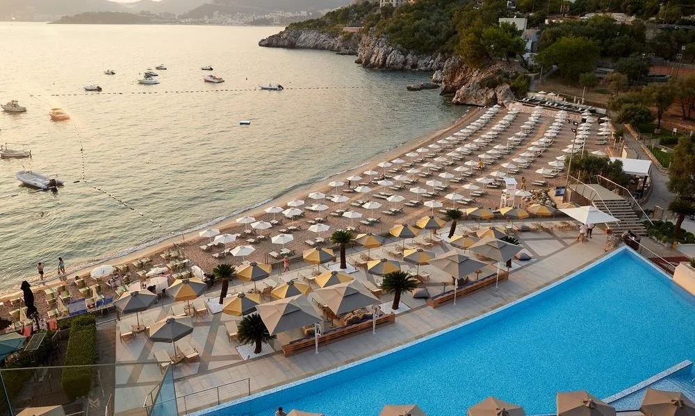Beach with rows of sun umbrellas and lounge chairs, a swimming pool, and boats anchored offshore in a cove.