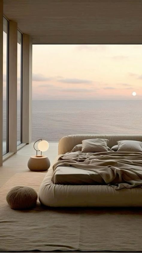 Modern bedroom with large floor-to-ceiling windows overlooking the ocean at sunset, featuring a low bed with neutral bedding, a small bedside table with a lamp, and a round cushion on the floor.