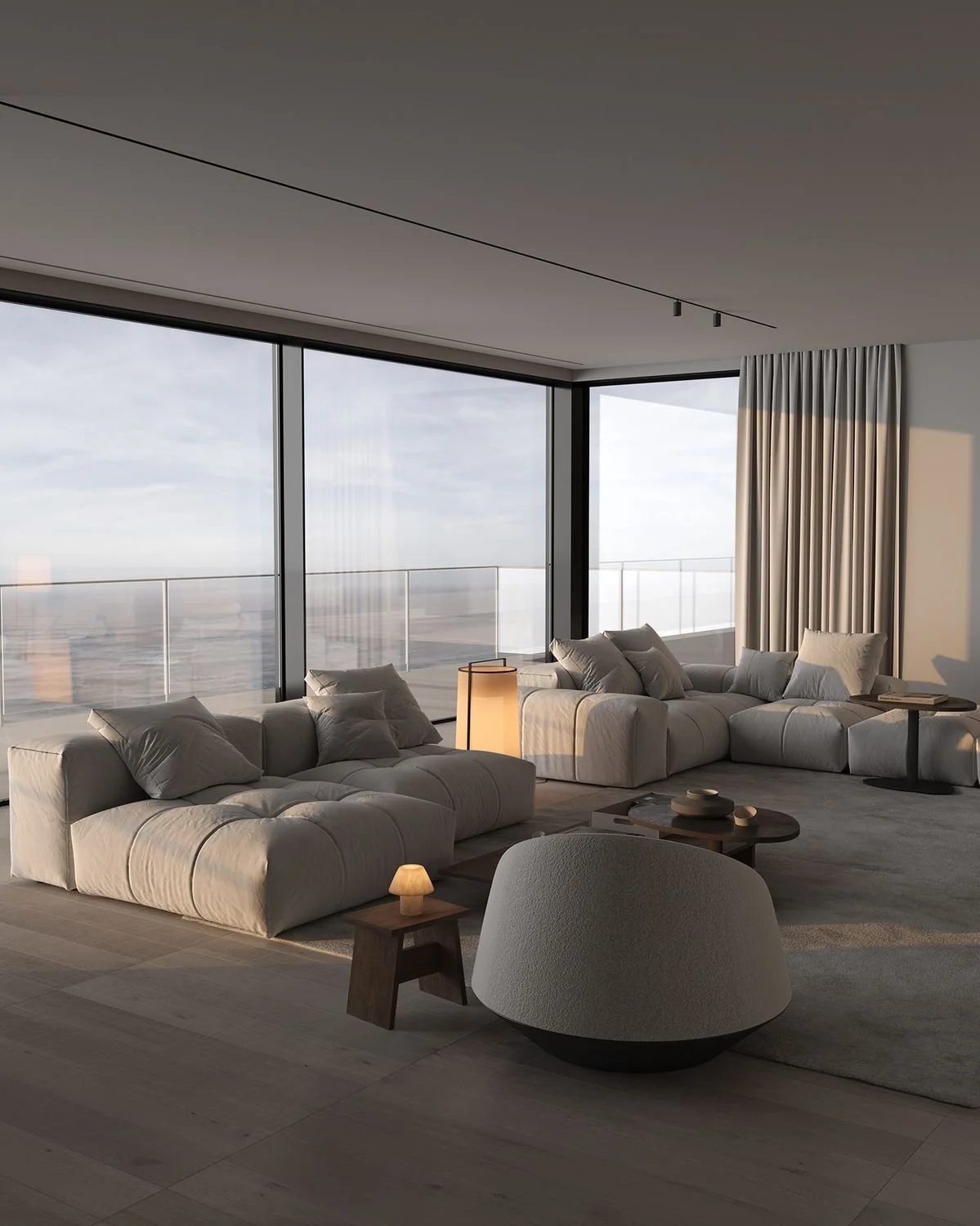 Modern living room with large floor-to-ceiling windows, white sofas with cushions, a wooden side table with a small lamp, a coffee table with decorative items, and beige curtains.