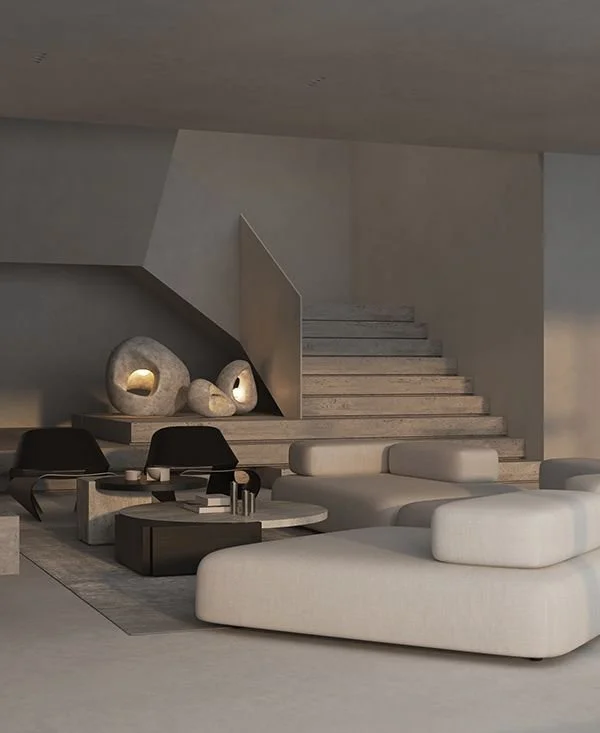 Modern living room with white sectional sofa, black chairs, round coffee tables, abstract stone sculptures with lit openings, and staircase against a minimalist gray wall.