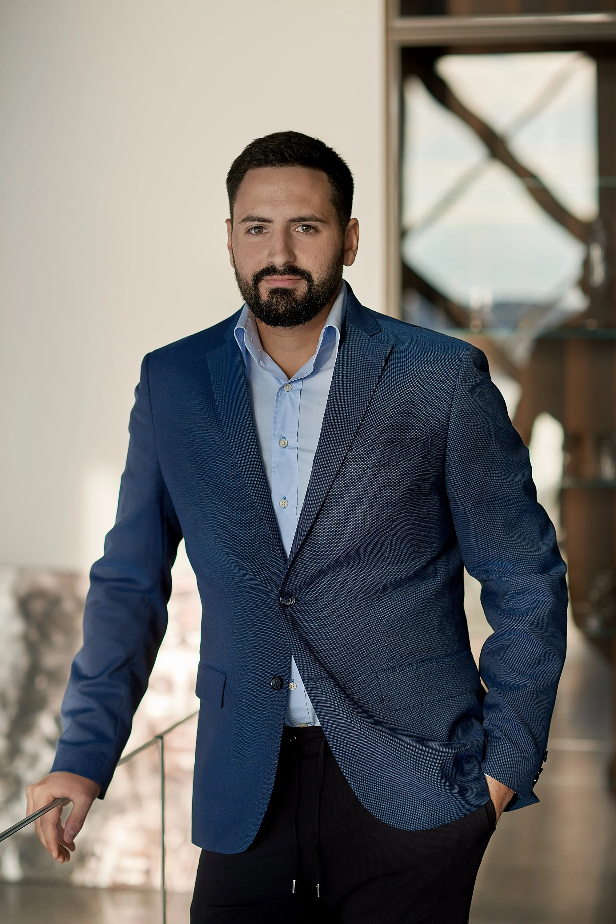 A man with dark hair and a beard wearing a blue suit and light blue shirt posing indoors near a glass railing with a blurred city view in the background.