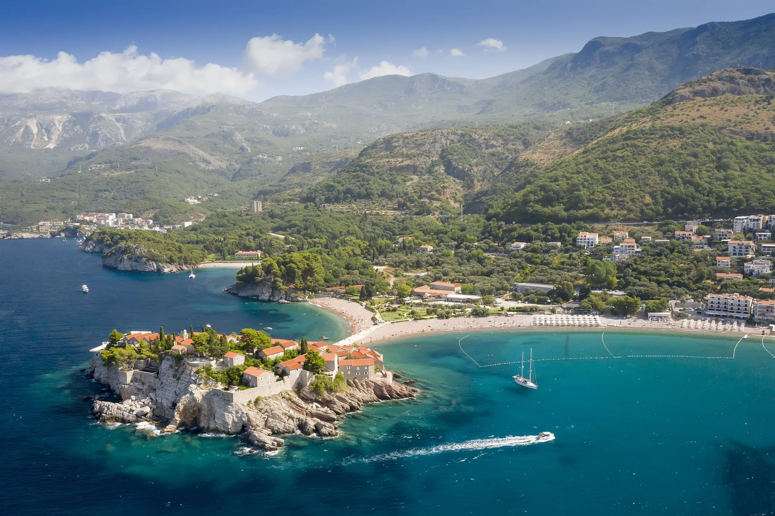Aerial view of a coastal area with a small island featuring buildings and red roofs, surrounded by turquoise water, with a sandy beach, boats, lush green hills, and mountains in the background under a partly cloudy sky.