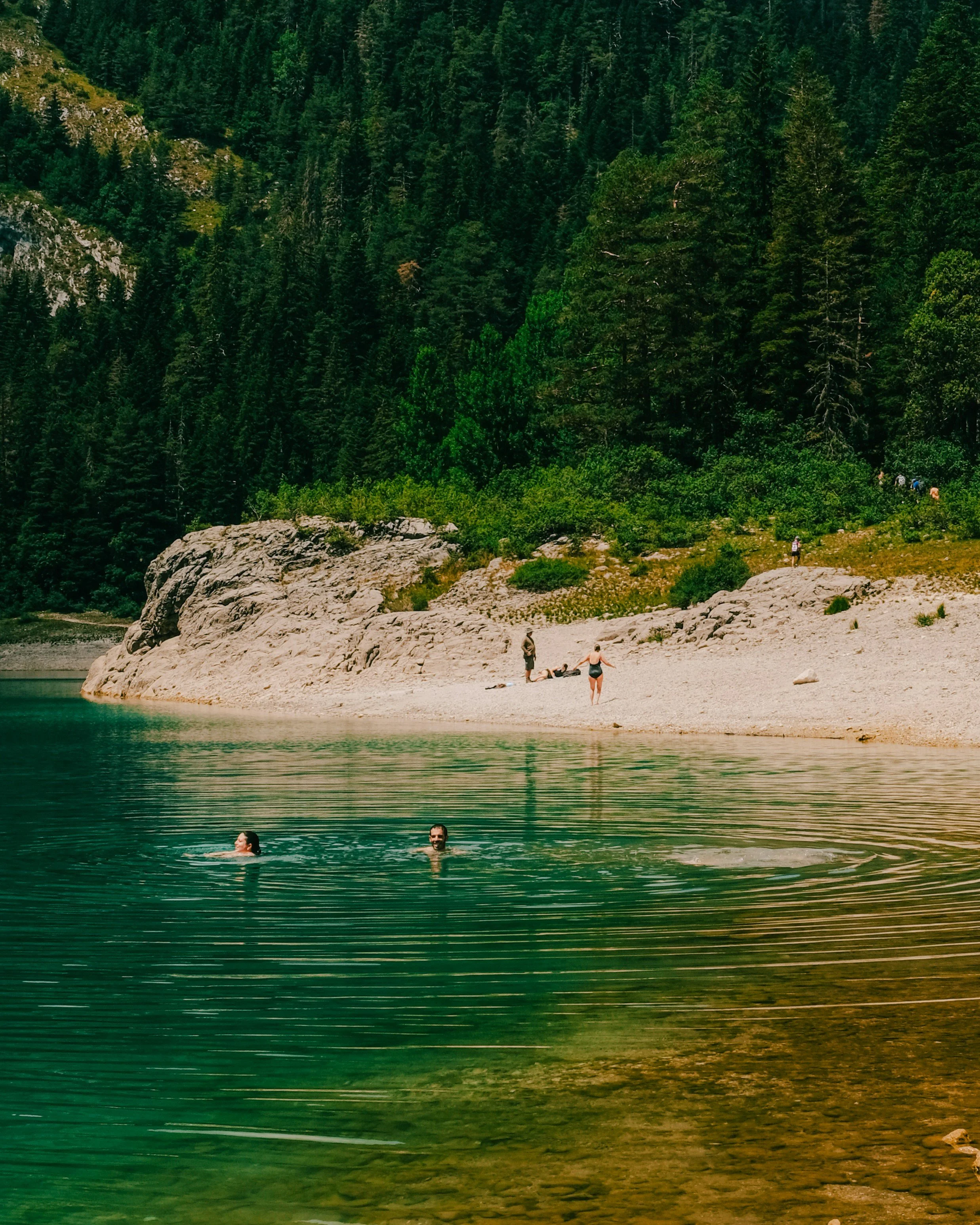 People swimming in a lake with a sandy shoreline, surrounded by green trees and rocky hills.