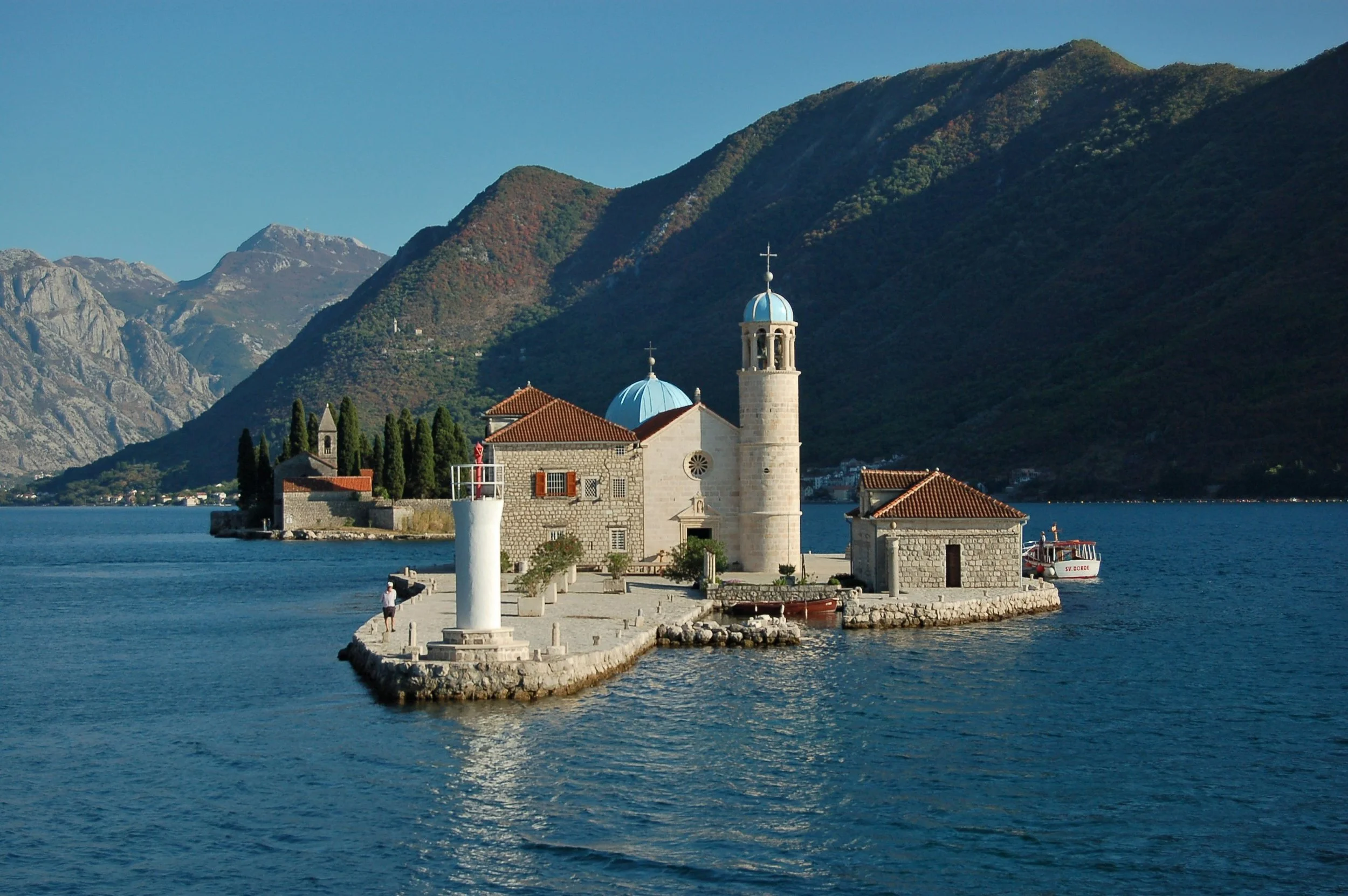 Small stone church on a pier with a lighthouse, surrounded by water, mountainous landscape in the background.