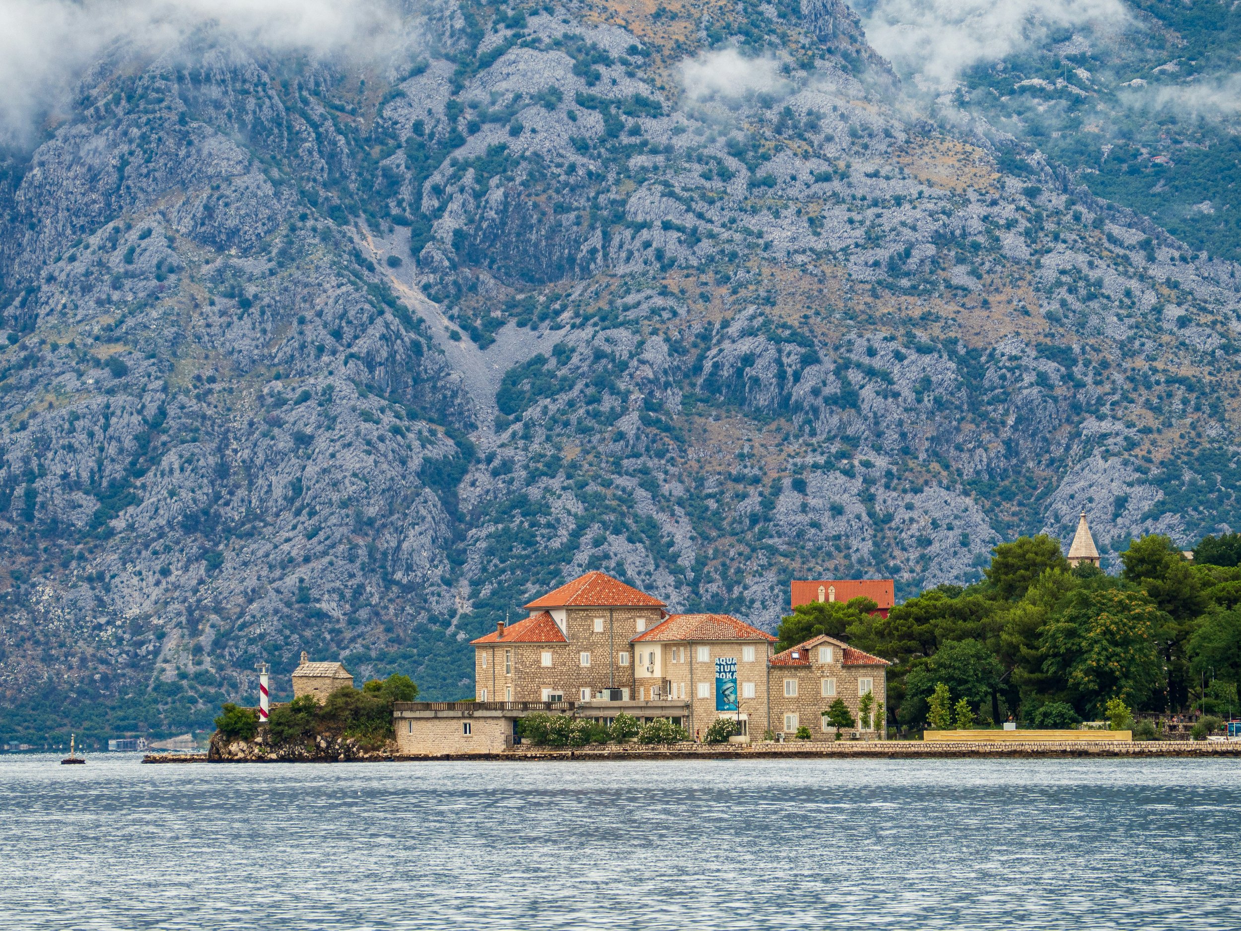 A small island with old stone buildings and red-tiled roofs, surrounded by water, with a mountainous landscape in the background.