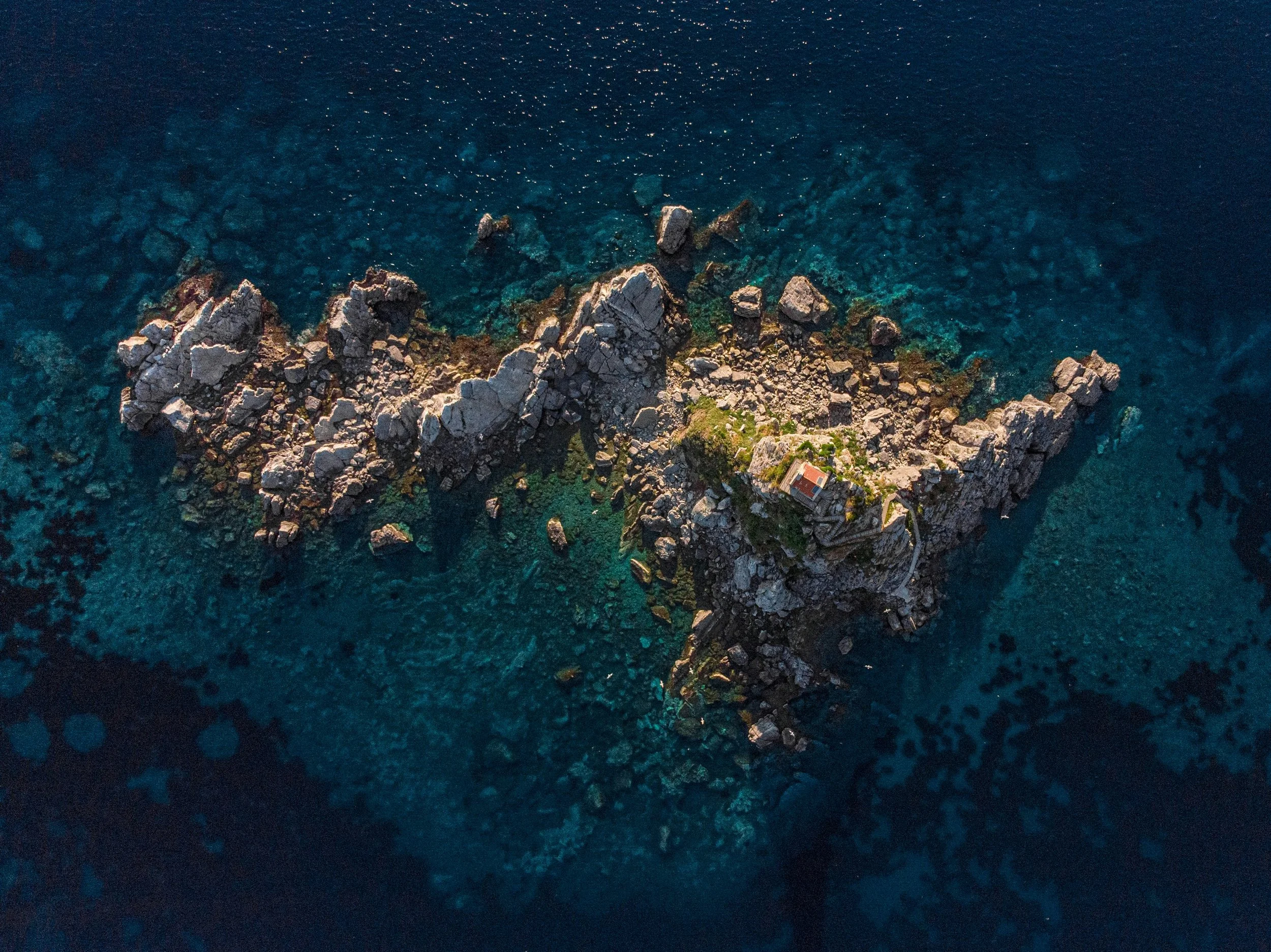 An aerial view of a rocky island with a single house on it, surrounded by clear blue ocean water.