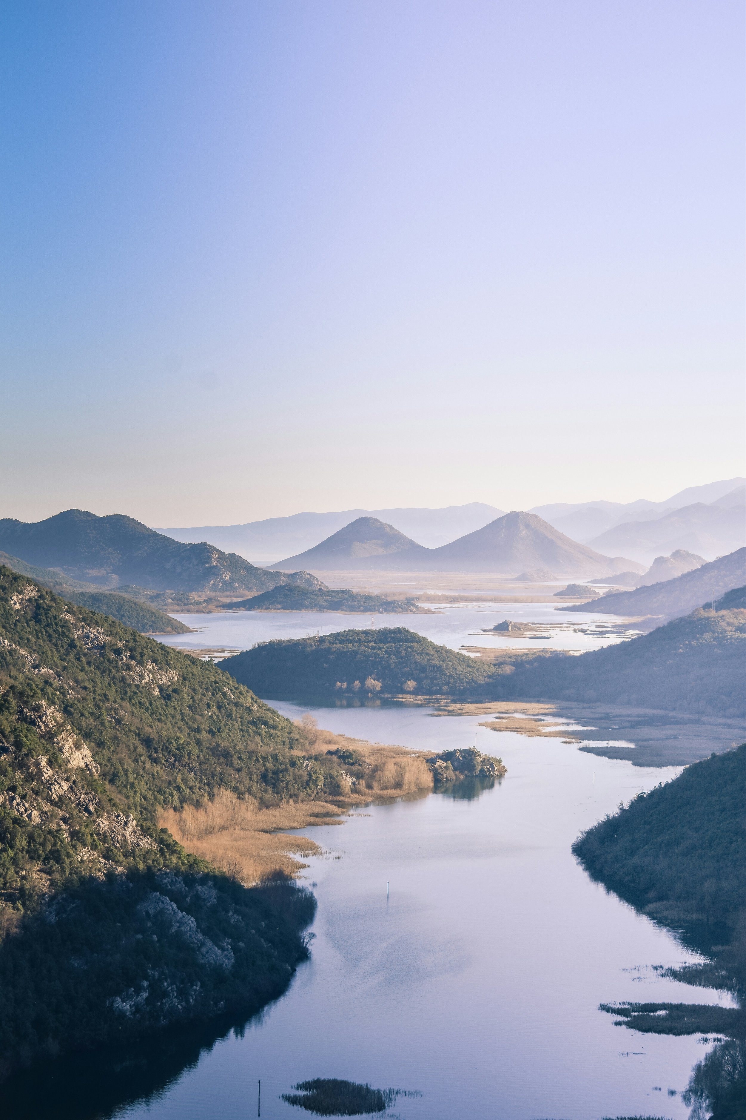 Scenic landscape of a river winding through lush green mountains with distant hills and mountains under a blue sky.