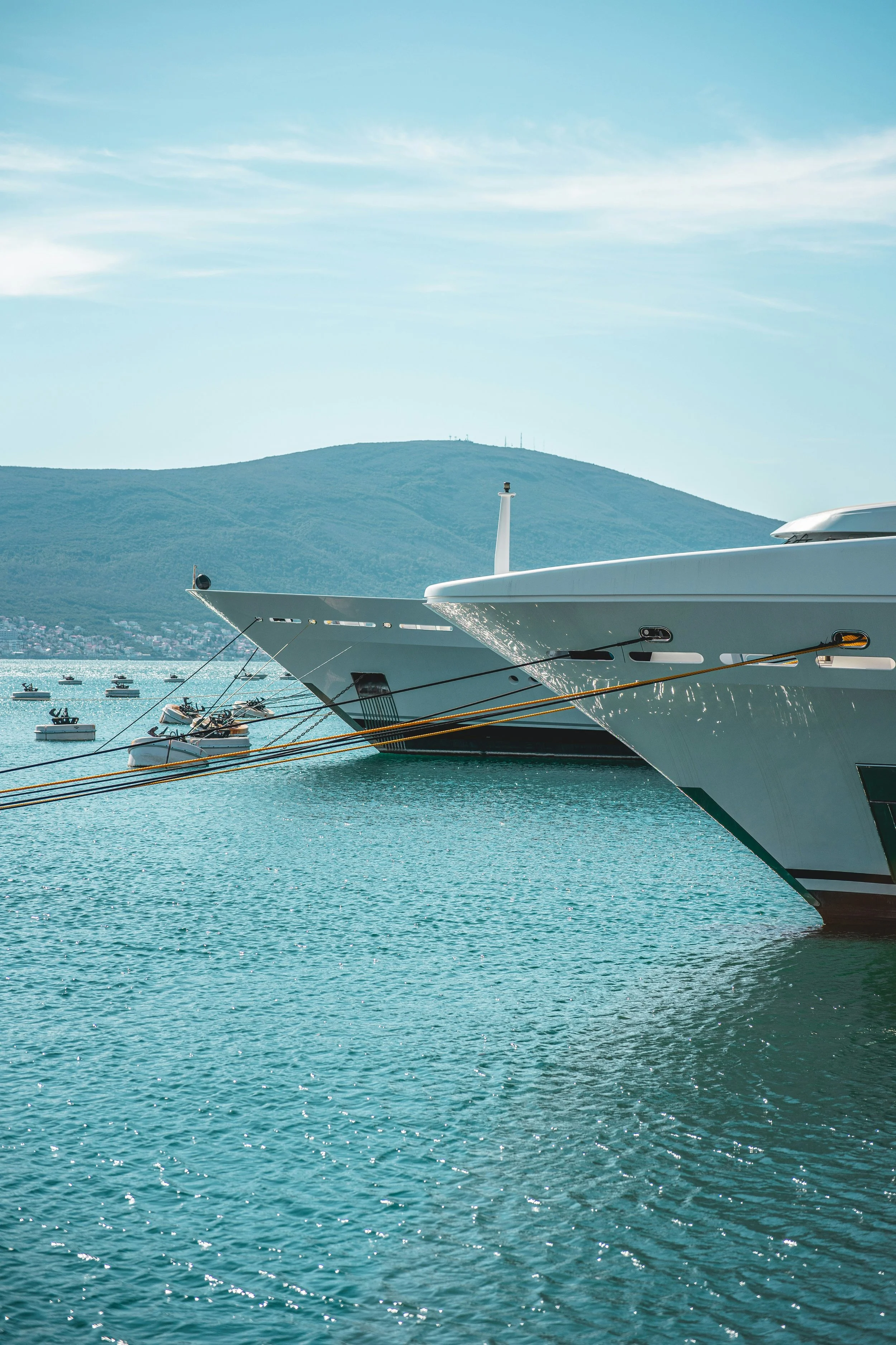 Two large yachts docked at a marina with smaller boats floating on the water and a mountain in the background under a partly cloudy sky.