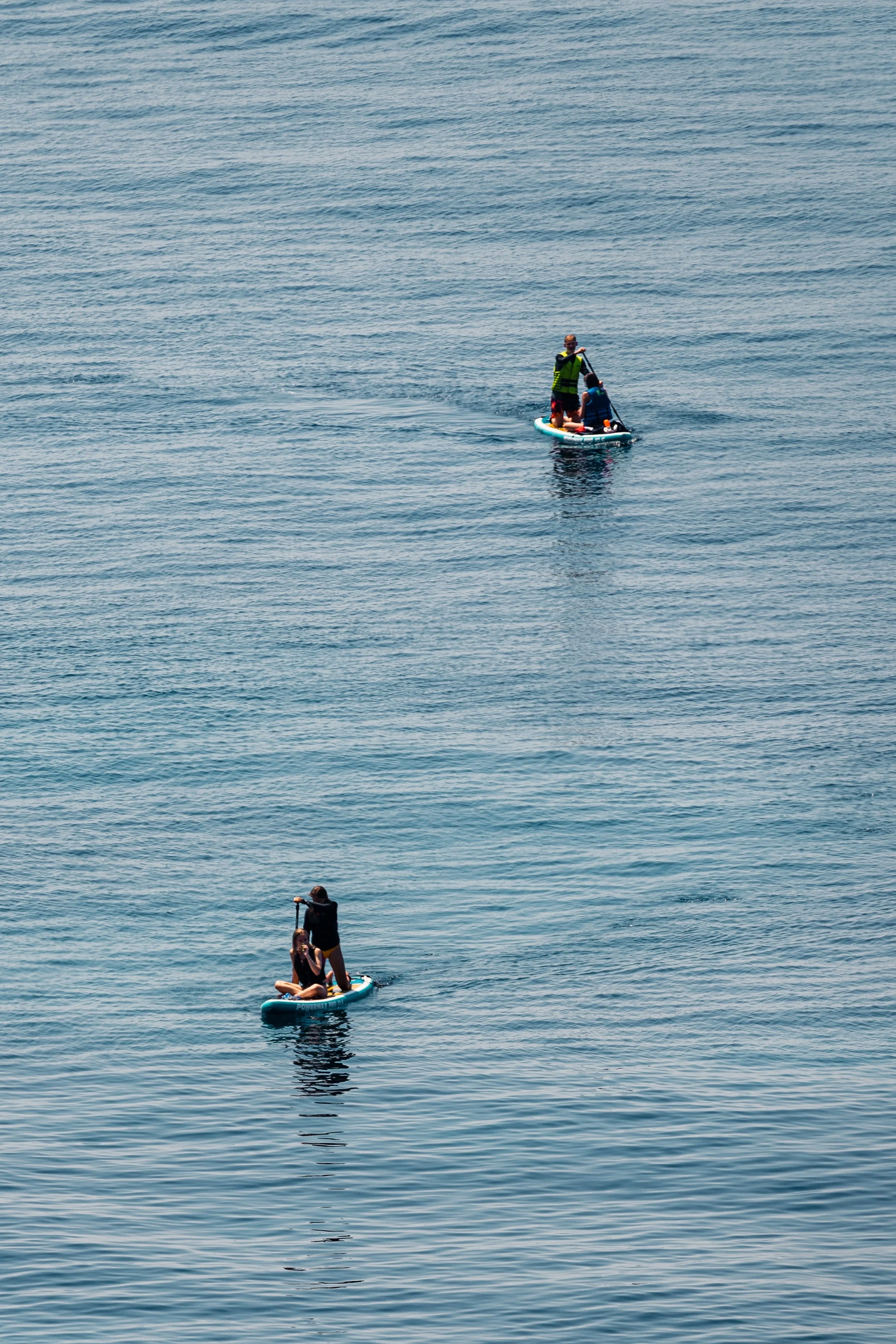 Two paddleboarders on the ocean, one standing with a paddle and the other sitting, both wearing life jackets.