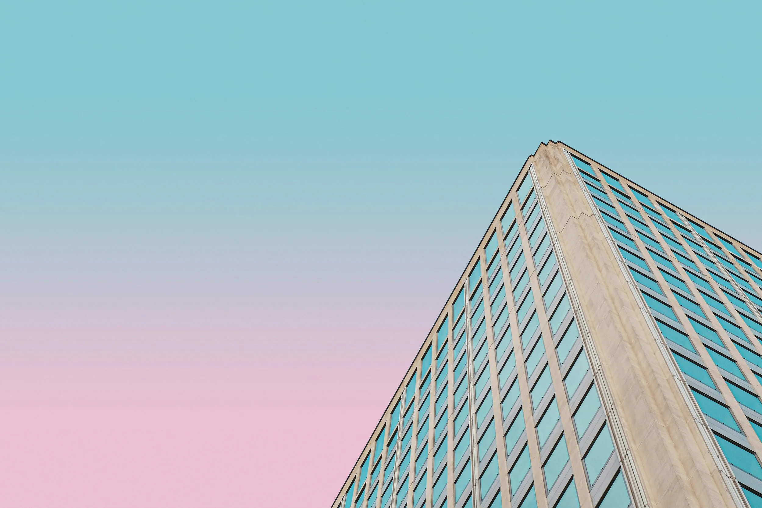 The corner of a tall modern office building with glass windows against a pastel-colored sky