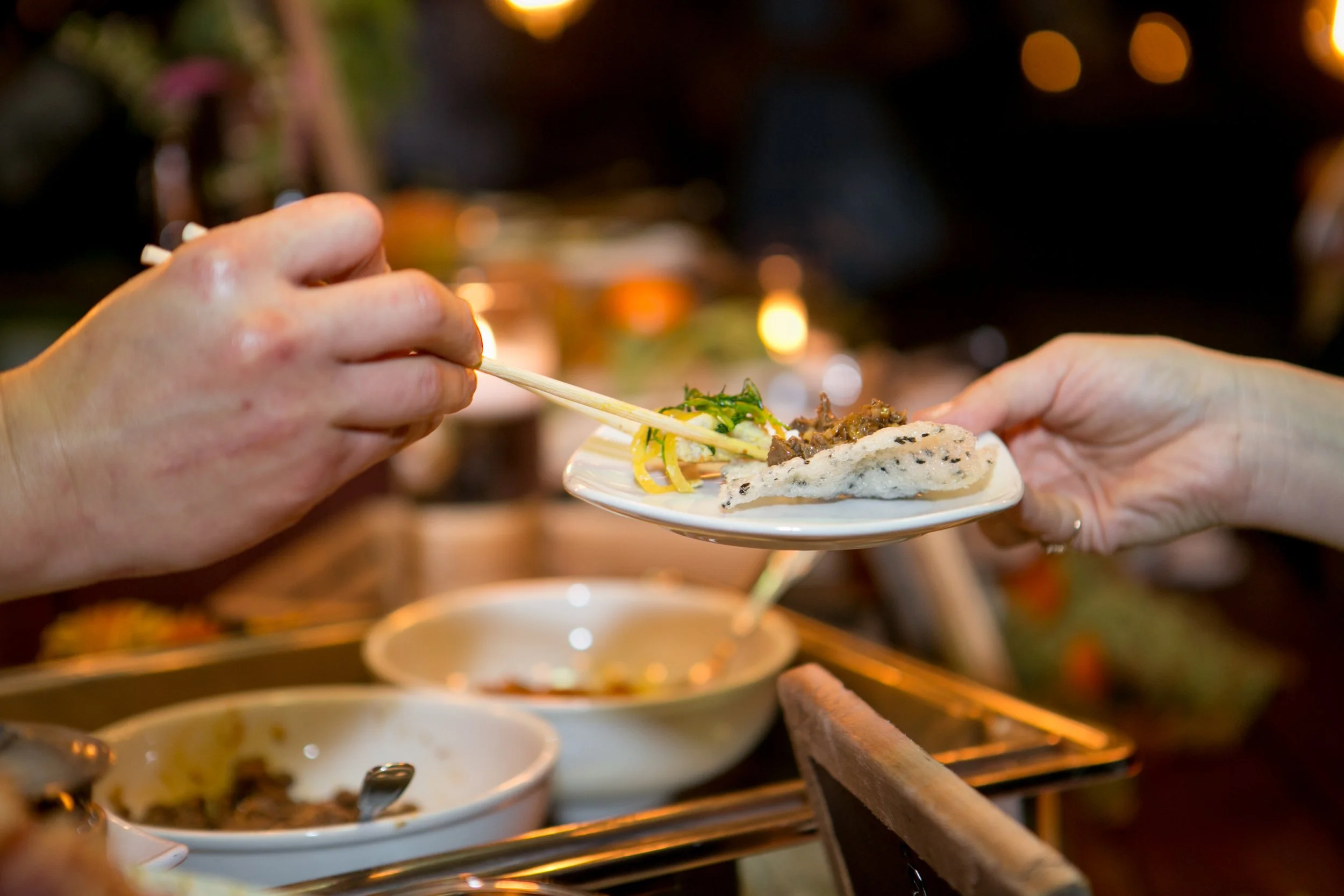 Two hands exchanging a small plate with Asian food, using chopsticks, in a warm, dimly lit setting with bowls of food in the background.