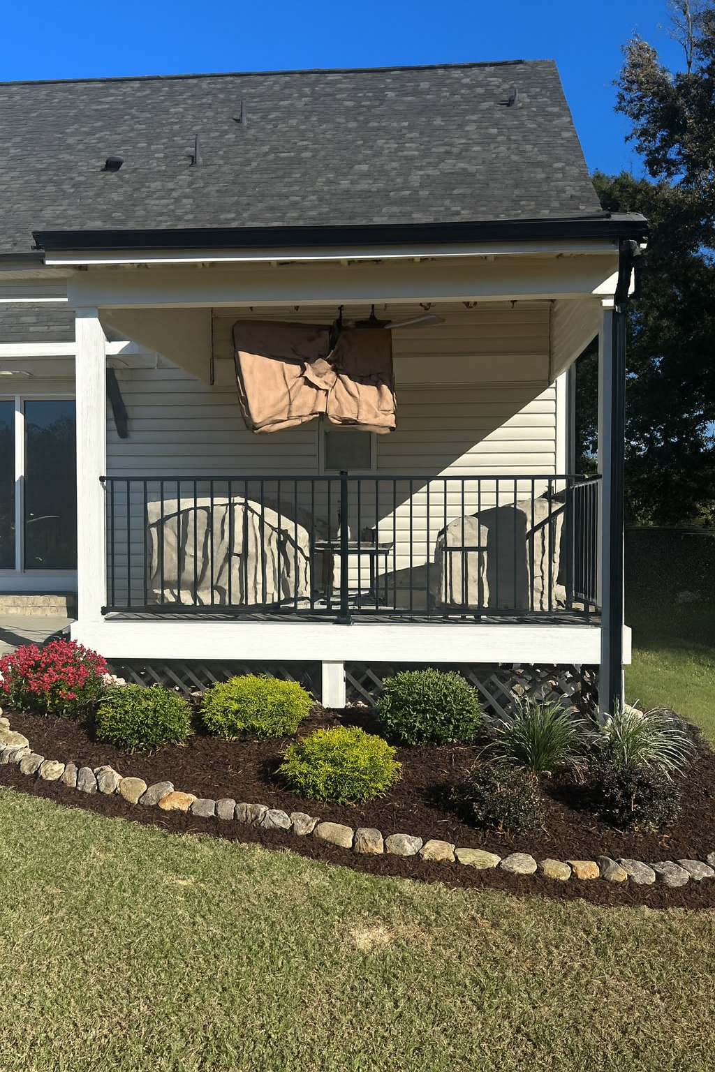 A house porch with a balcony, a ceiling fan, and an outdoor television covered with a brown tarp. The porch has a black railing, and there are large rocks behind the railing. In front of the porch, there is a flower bed with flowers and shrubs, bordered by small rocks, and a lawn.
