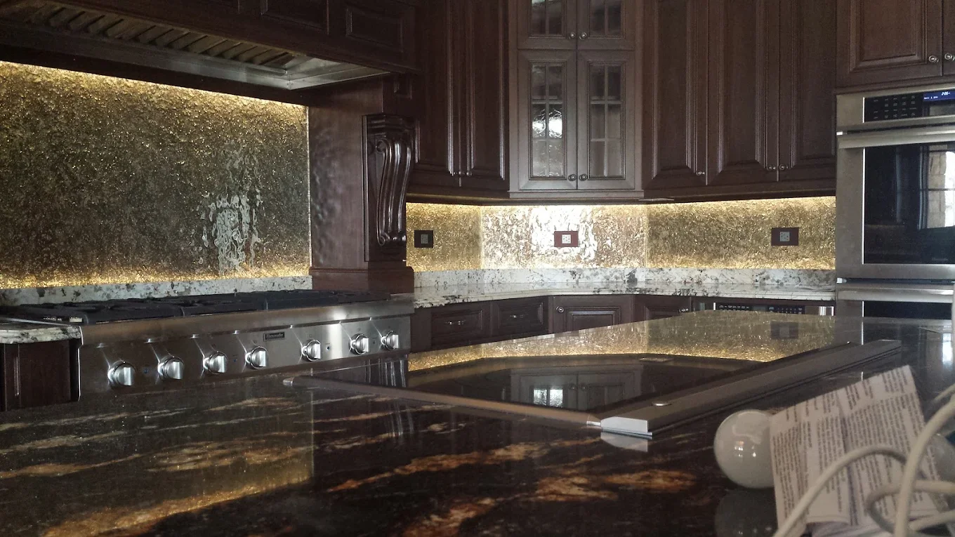 A luxurious kitchen with dark wooden cabinets, a granite countertop, and a gold-toned backsplash. There is a stainless steel stove on the left, a double oven on the right, and some items on the counter.