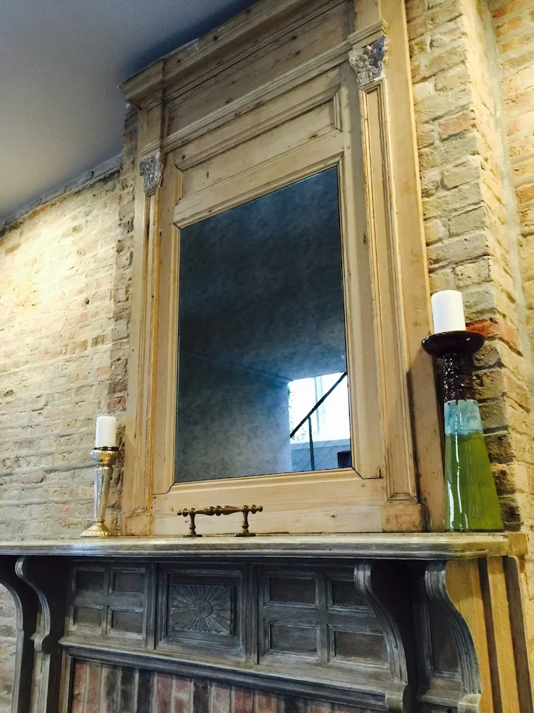 A large ornate mirror with a wooden frame mounted on a brick wall, with a marble-topped mantel below it. Two candle holders with white candles are placed on either side of the mirror on the mantel, and a decorative glass vase is on the right.