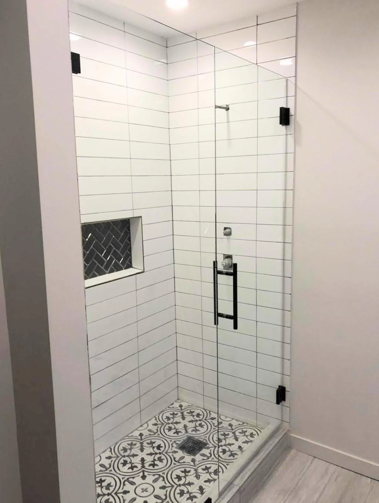 A modern walk-in shower with white rectangular subway tiles on the walls, black metal hardware, a small recessed niche with black and white patterned tiles, and a decorative black and white patterned floor, enclosed by clear glass panels.