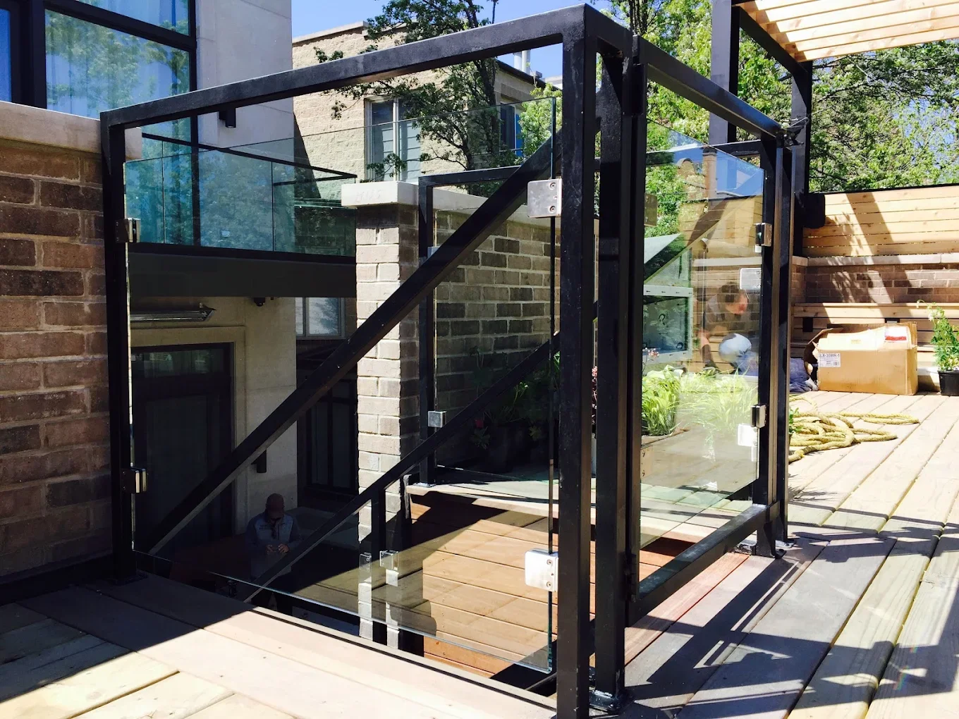 Outdoor wooden deck with glass railing and a modern black metal staircase leading below, surrounded by brick walls, greenery, and furniture.