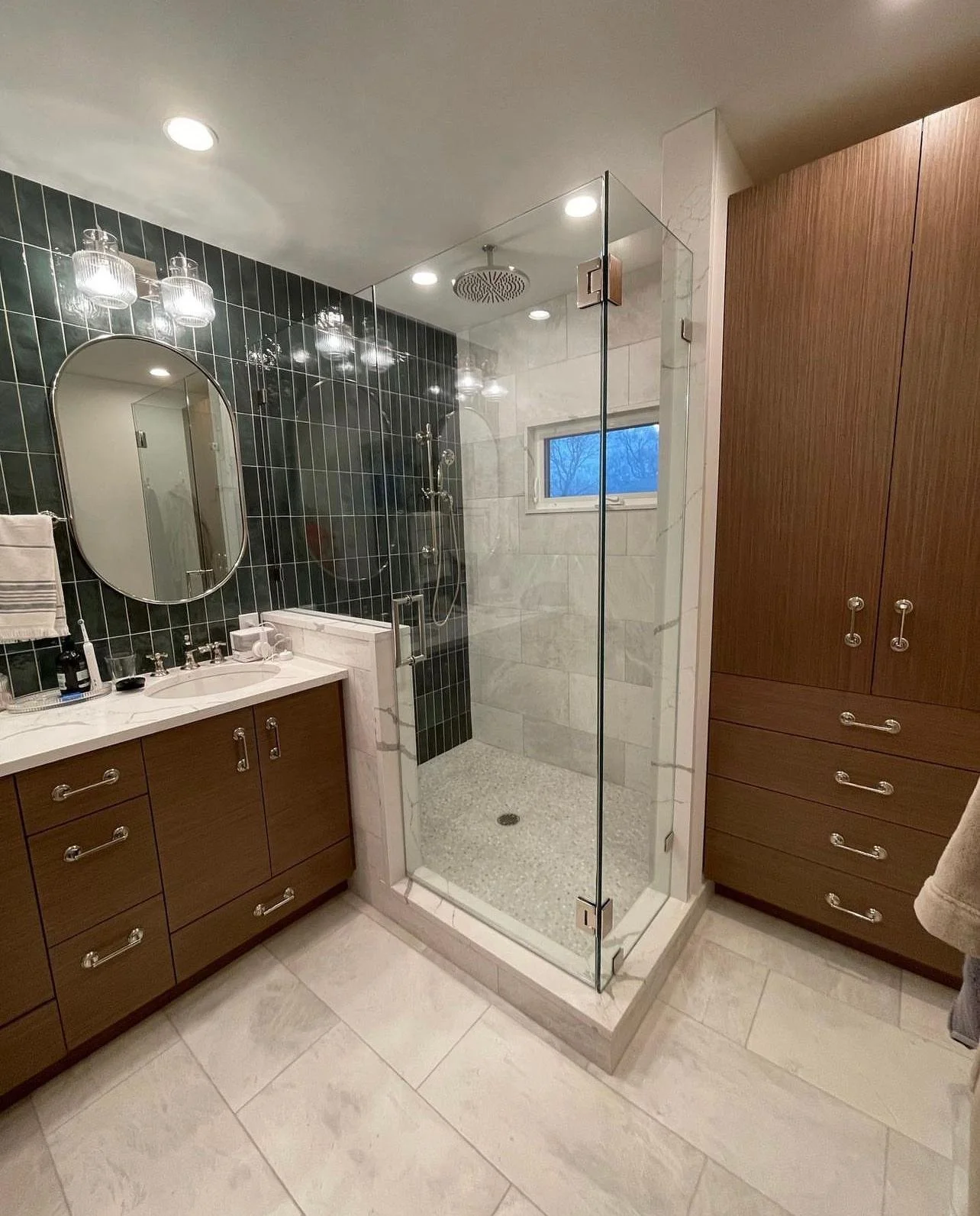 Modern bathroom with a glass shower enclosure, dark green tiled accent wall, white marble vanity with oval mirror, wooden cabinets, and beige floor tiles.