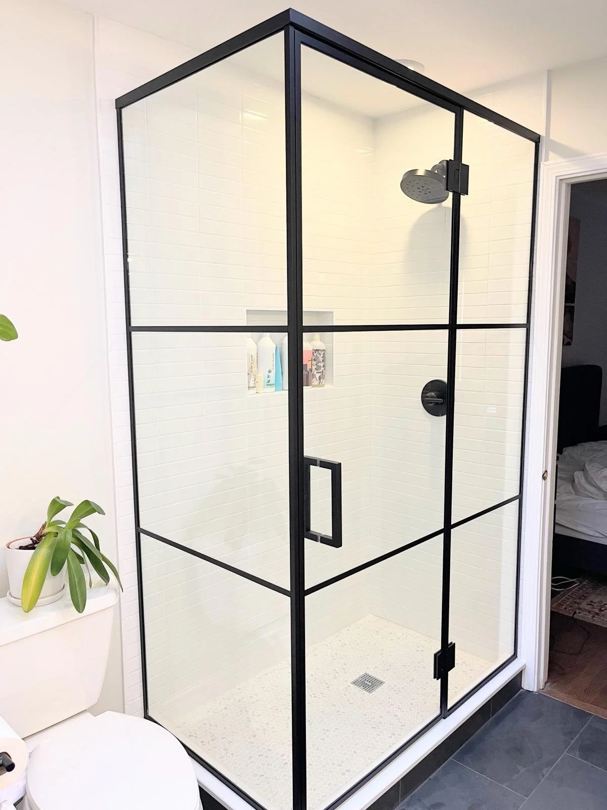 Modern glass shower enclosure with black framing and handle, white tiled interior, and a ceiling-mounted shower head, in a bathroom with white walls, a toilet, a potted plant, and adjacent to a room with a bed.