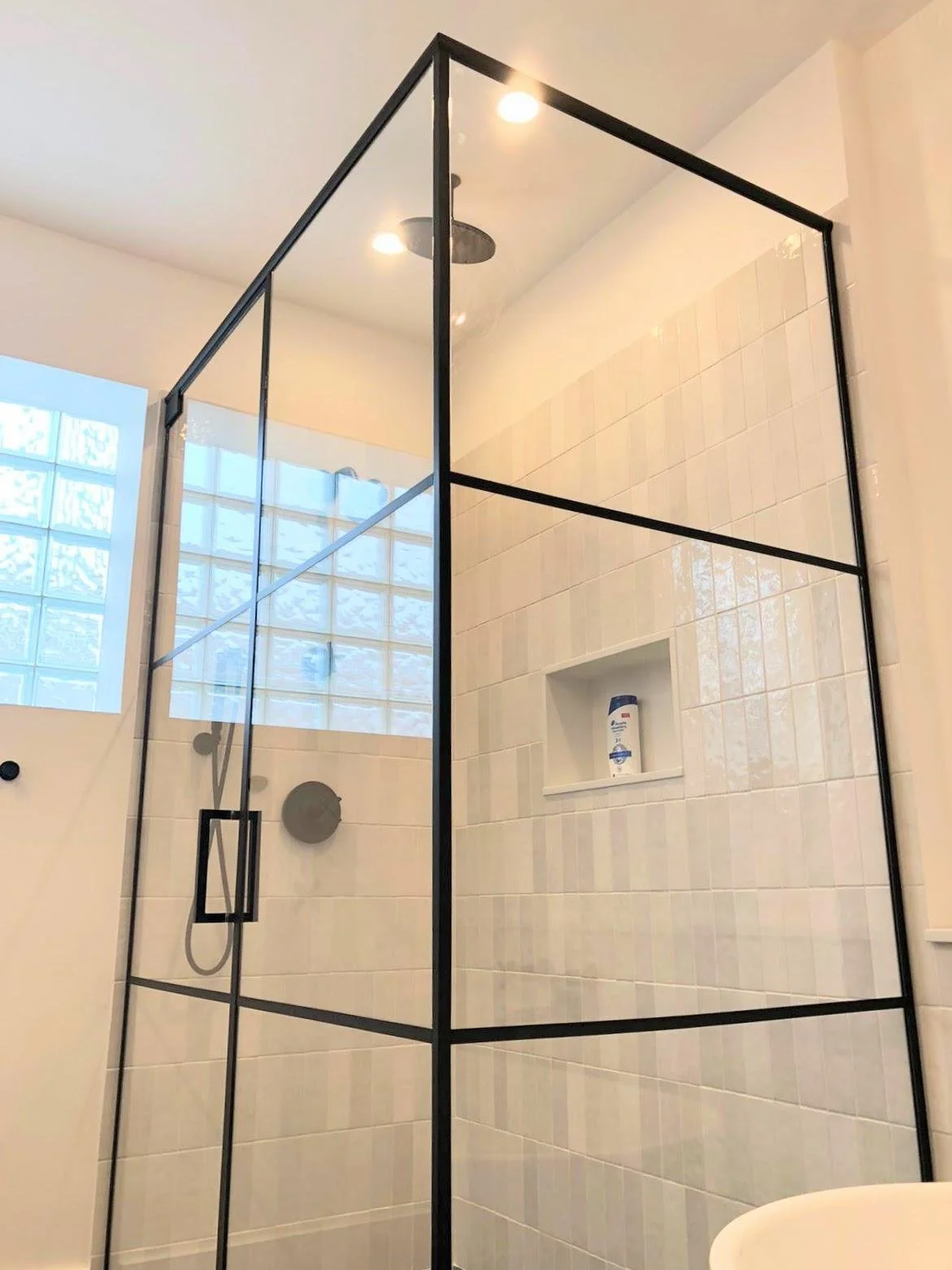 Modern bathroom shower with glass walls and black metal frame, white tiled walls, built-in niche with toiletries, ceiling-mounted showerhead, and window with frosted glass blocks.