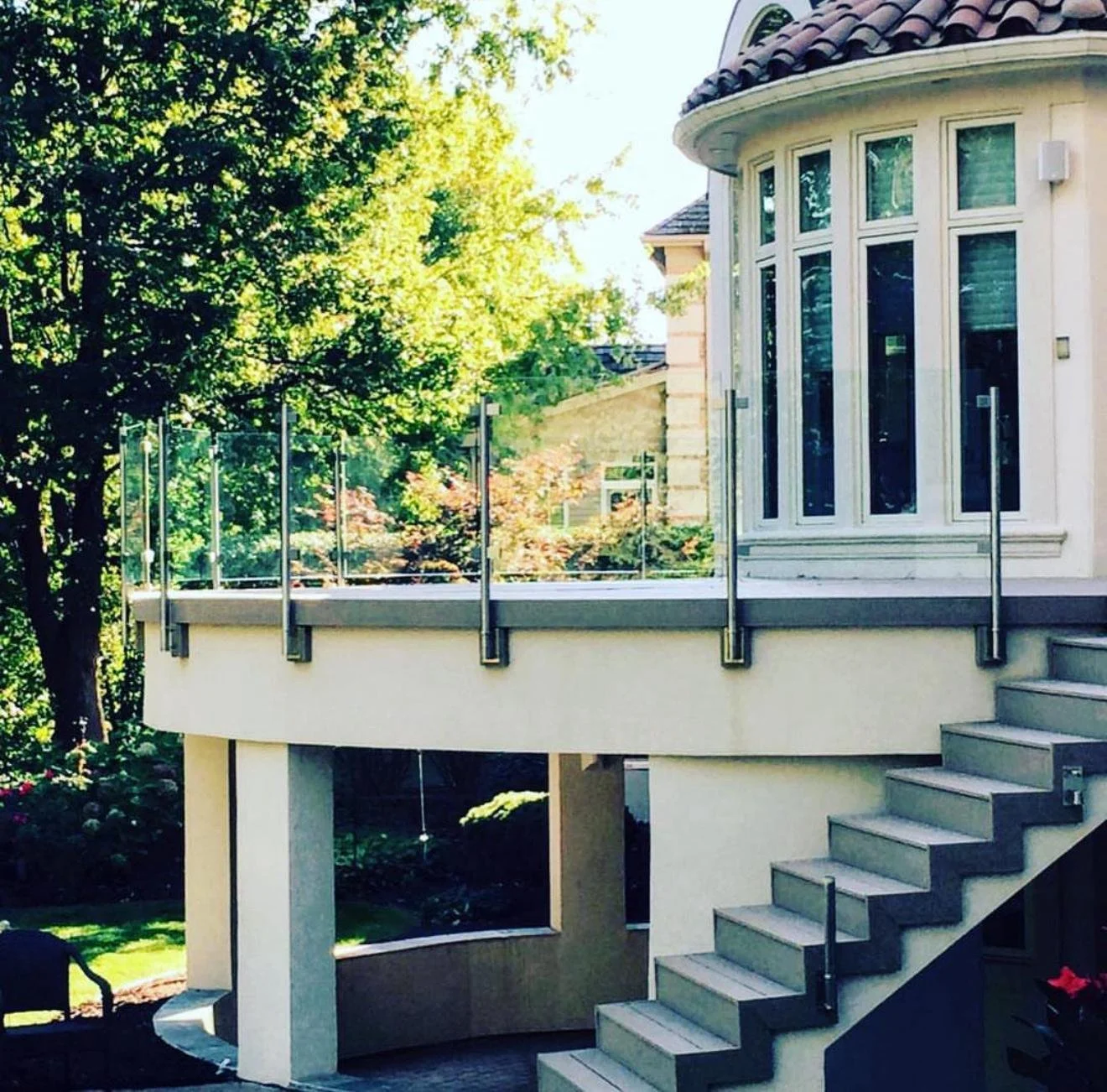 A white house with a rounded bay window, an outdoor staircase, and a glass railing on a balcony, surrounded by trees with green foliage.