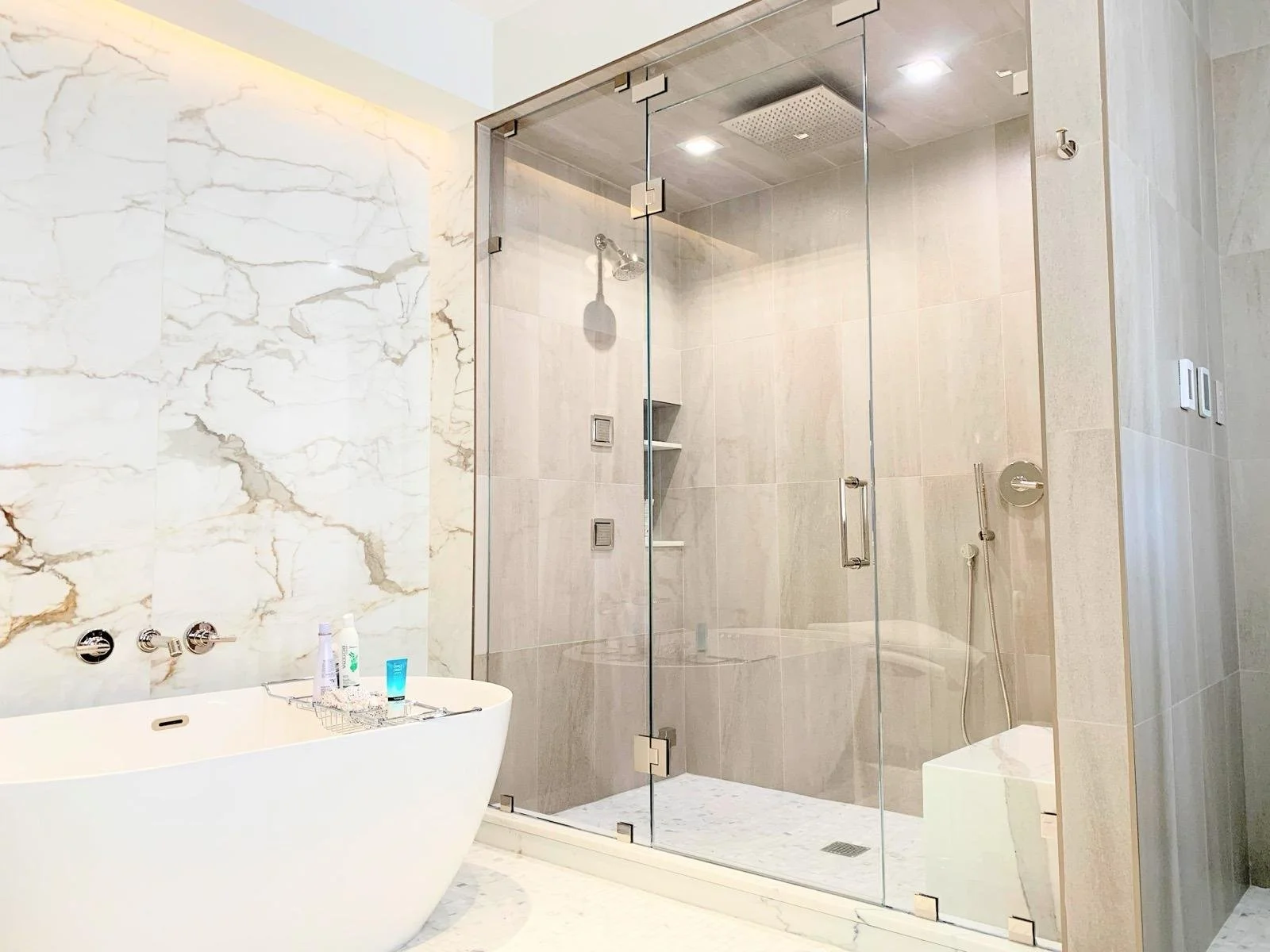Modern bathroom with a white freestanding bathtub, marble walls, and a glass-enclosed shower with rainfall showerhead and handheld sprayer.