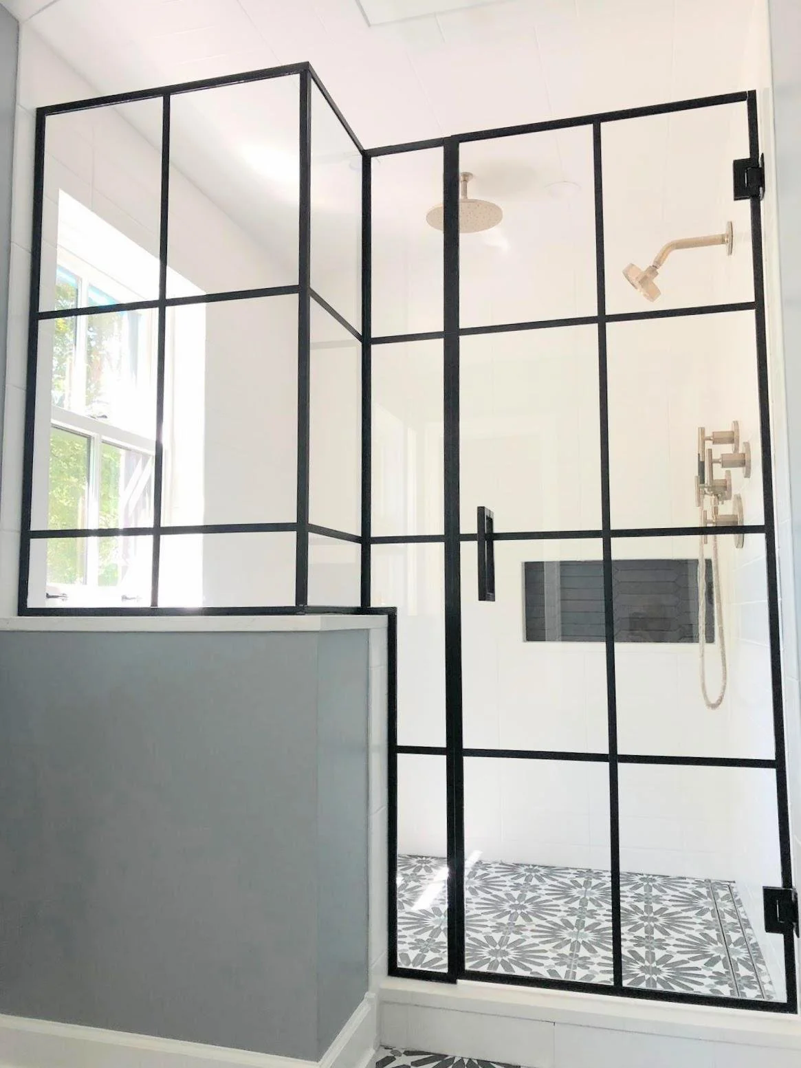 Modern walk-in shower with black metal framing, glass door, white tiled walls, patterned floor tiles, and ceiling-mounted showerheads.