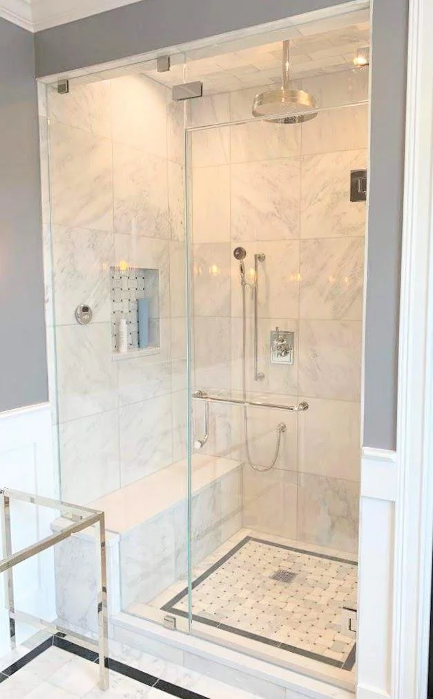 A modern walk-in shower with marble tiles, a glass enclosure, a rain shower head, built-in niche with toiletries, and safety grab bars.