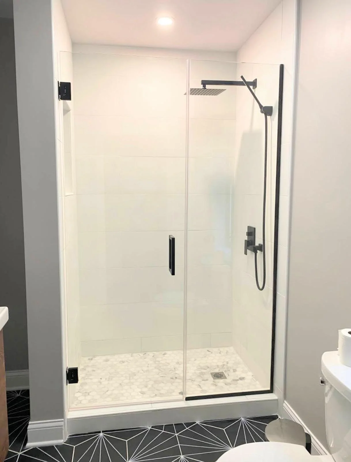 Modern walk-in shower with glass doors, black fixtures, and white tiled walls, in a bathroom with black and white geometric patterned flooring.