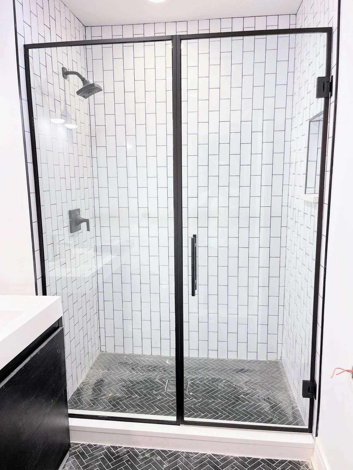 A modern glass shower enclosure with black metal framing, white subway tile walls, a black herringbone tile floor, and a rain showerhead.