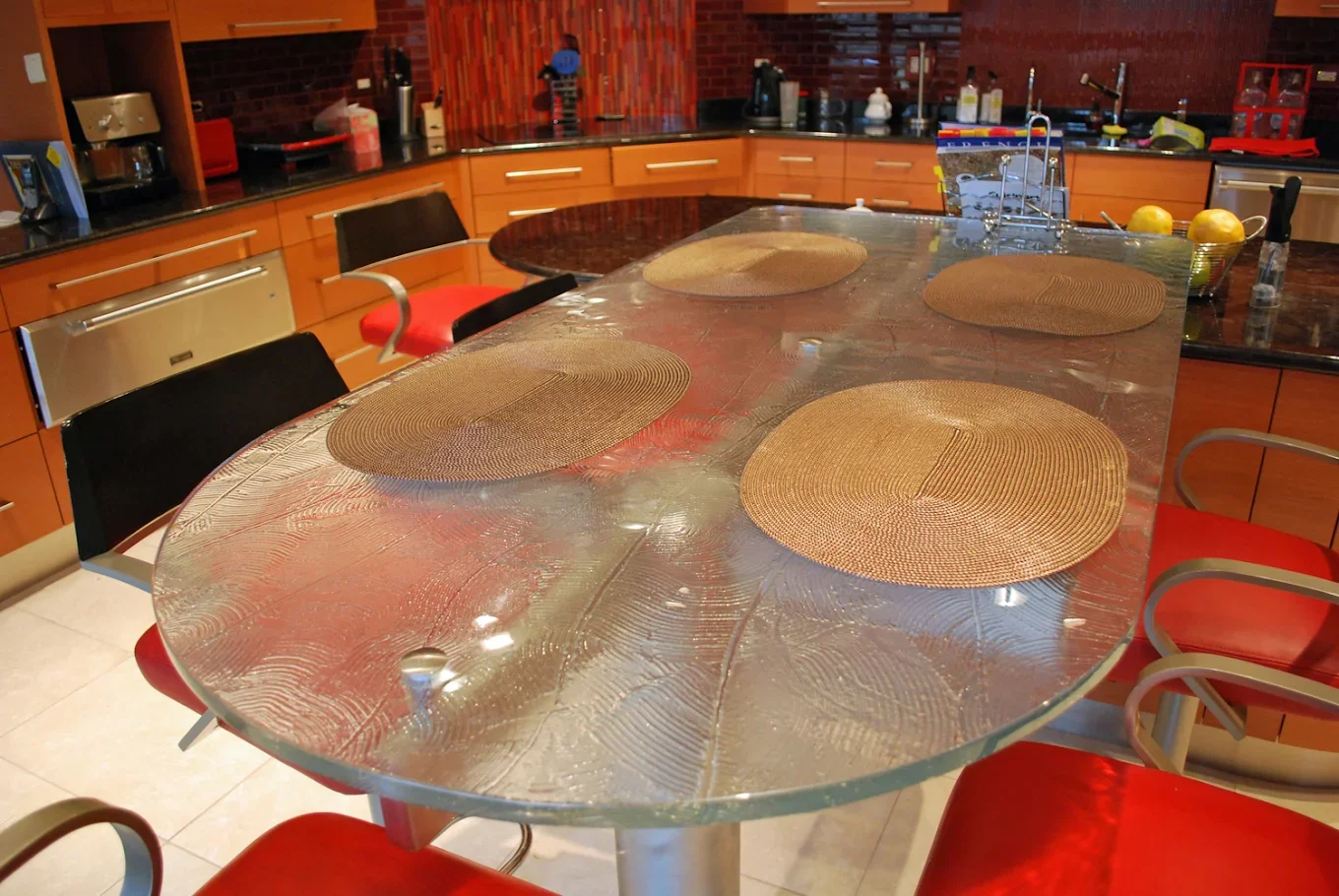 A glass dining table with four round woven placemats and red cushioned chairs in a kitchen with wooden cabinets and a black countertop.
