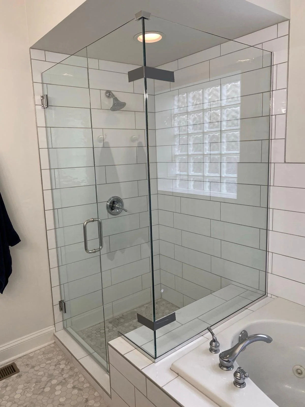 A glass-enclosed shower with white subway tiles and a small window with glass blocks, next to a bathtub with chrome fixtures.