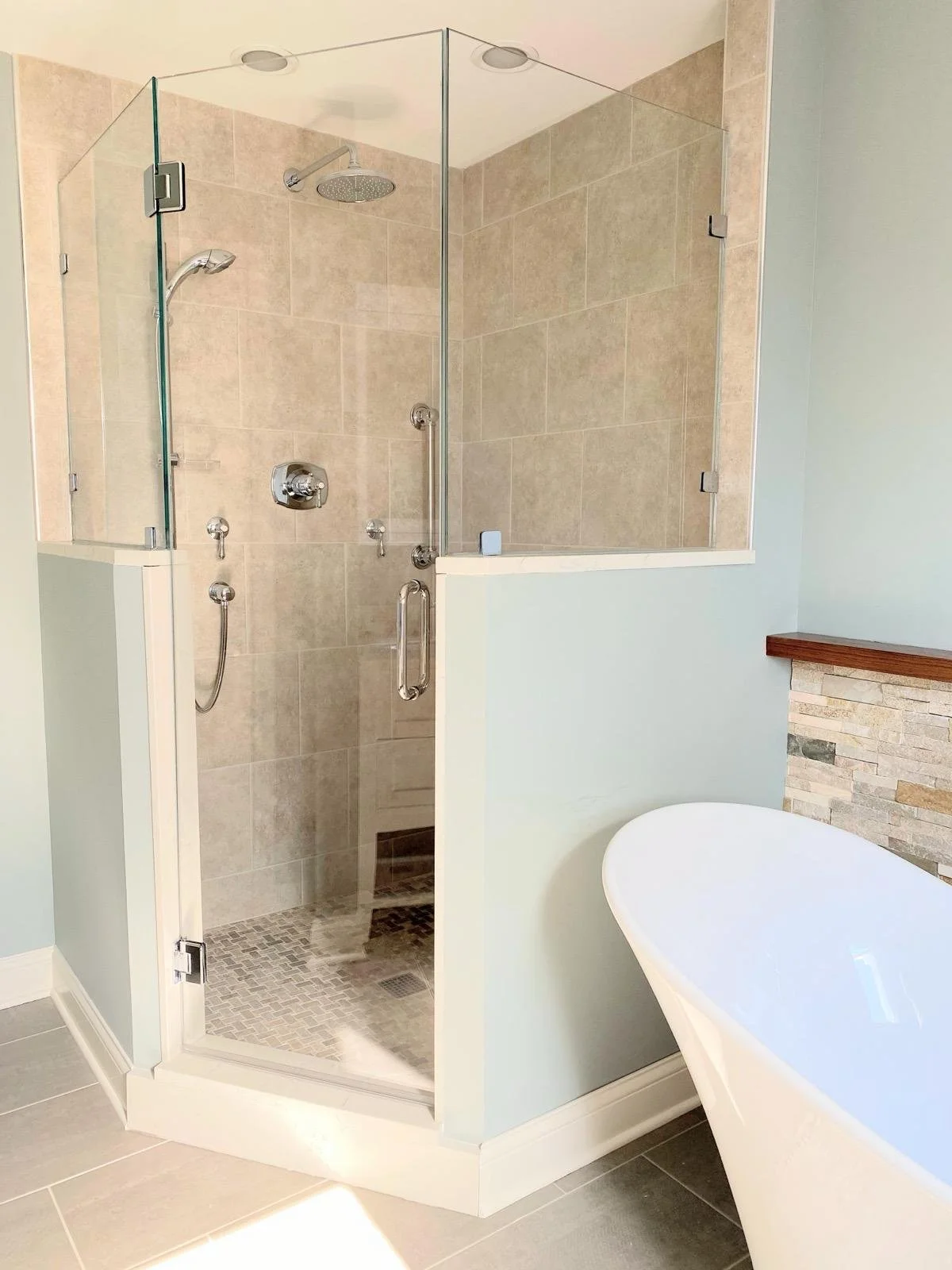 A modern bathroom with a glass-enclosed shower featuring beige tile walls and a tiled shower floor, and a white freestanding bathtub beside a partial wall with a stone accent.