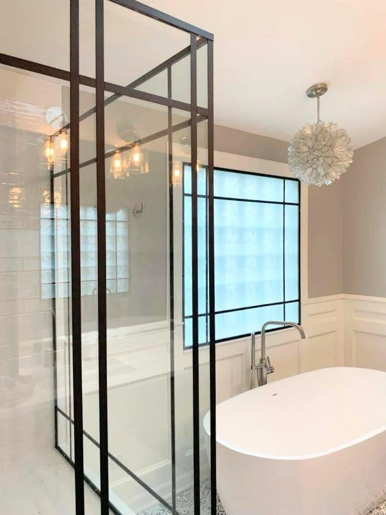 Bathroom with a standalone bathtub, large window with frosted glass, wall-mounted faucet, and a glass-enclosed shower with black metal frame. The room has light-colored walls, white wainscoting, and a modern white pendant light fixture.