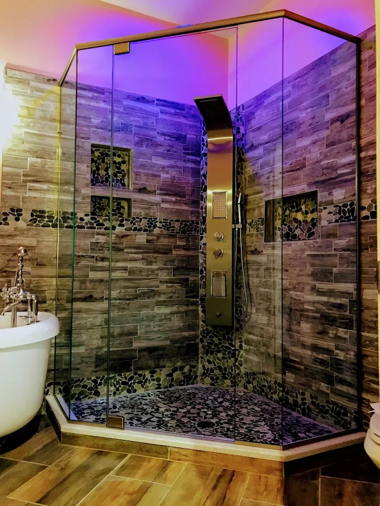 A modern bathroom shower with glass enclosure, wood-look tile walls, pebble tile floor, built-in shelves, and multicolored purple and pink lighting.