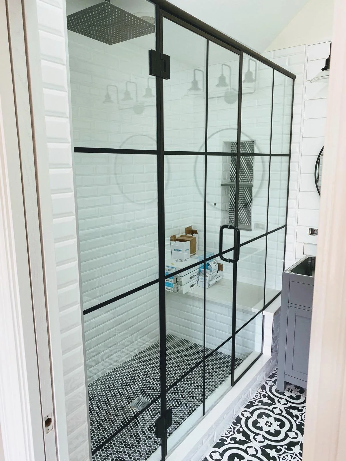 A modern walk-in shower with black-framed glass doors, a rain showerhead, white subway tile walls, and black and white patterned floor tiles in a bathroom.