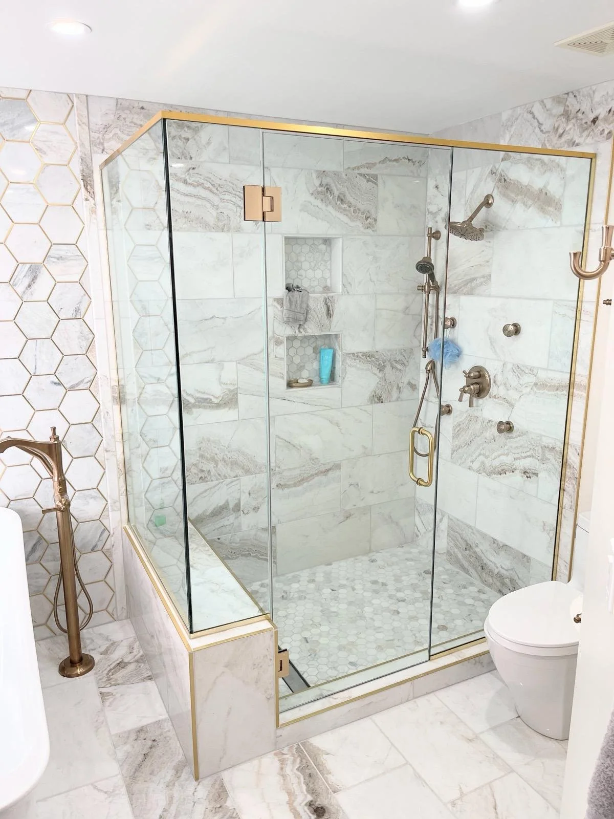 Modern bathroom with glass-enclosed shower featuring marble tiles, built-in shelves, and gold accents, alongside a white toilet and a golden towel rack.