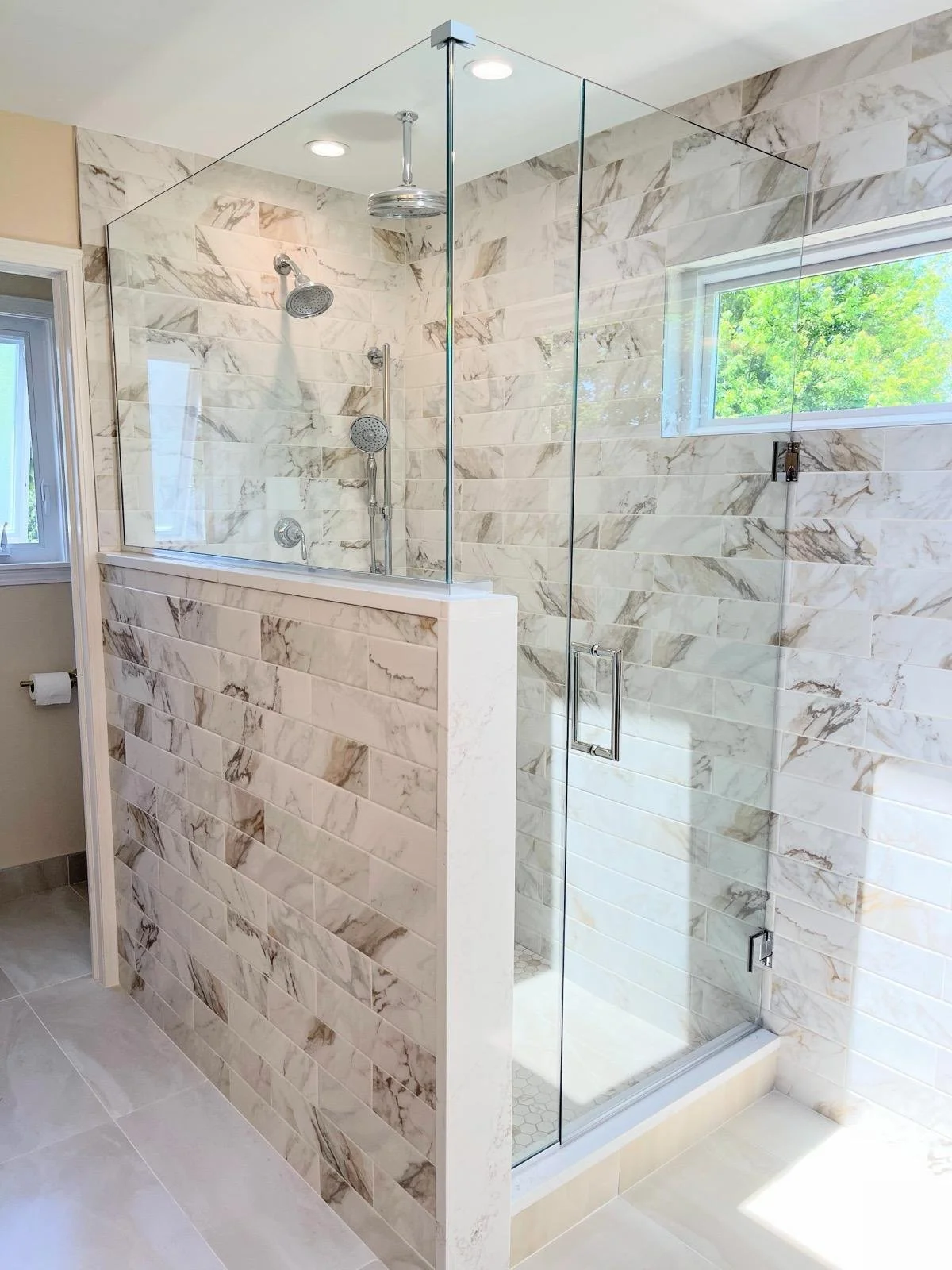 Bathroom shower with clear glass enclosure, marble tile walls, and a window allowing natural light, with two showerheads and a rain shower fixture.