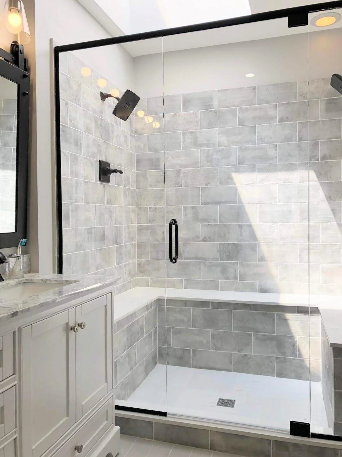 A modern shower with a glass door, black fixtures, gray tile walls, and a built-in bench.