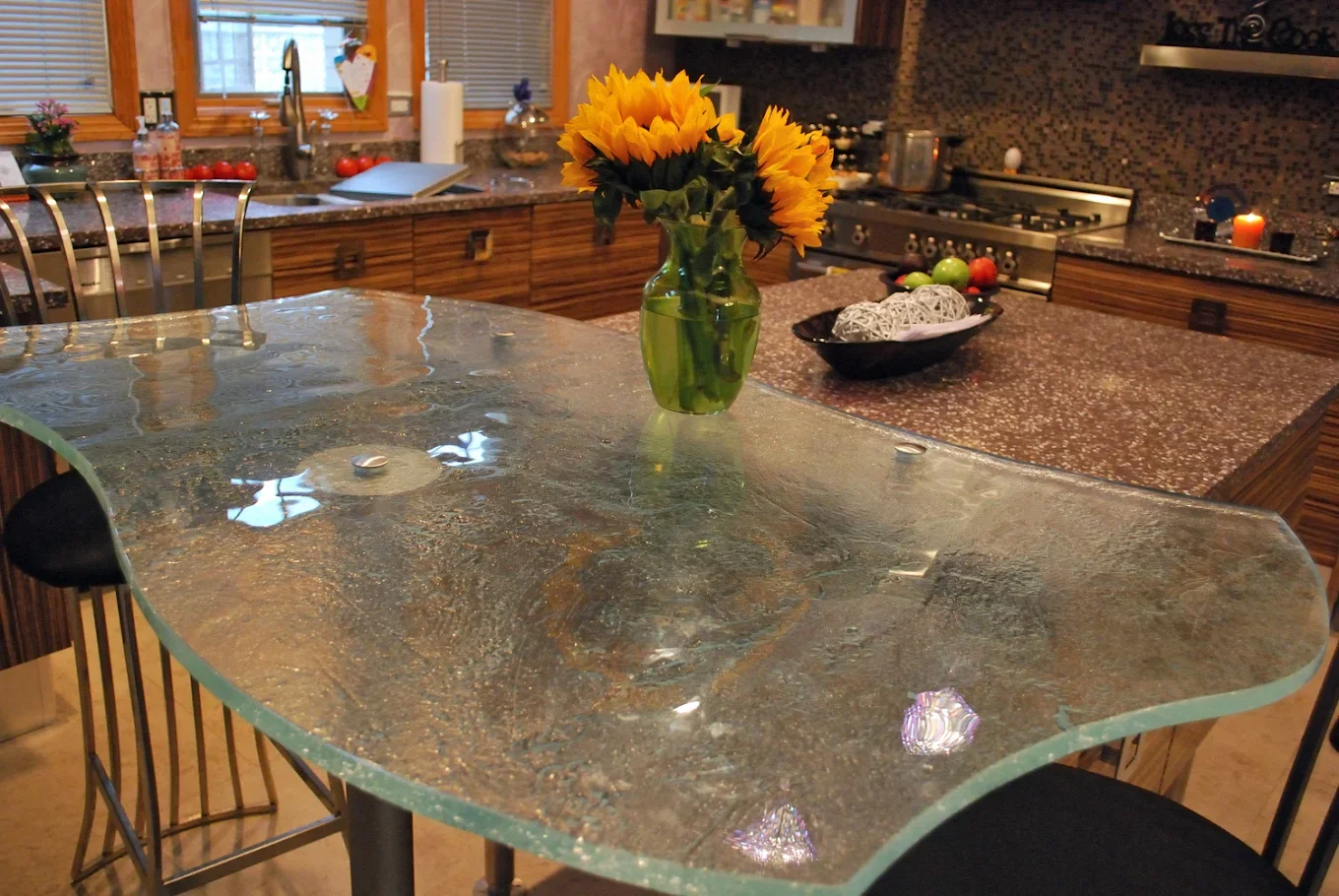 A kitchen with a glass table, a vase of sunflowers, and dark countertops. There are various kitchen items, including a bowl of apples, a loaf of bread, and a lit candle.