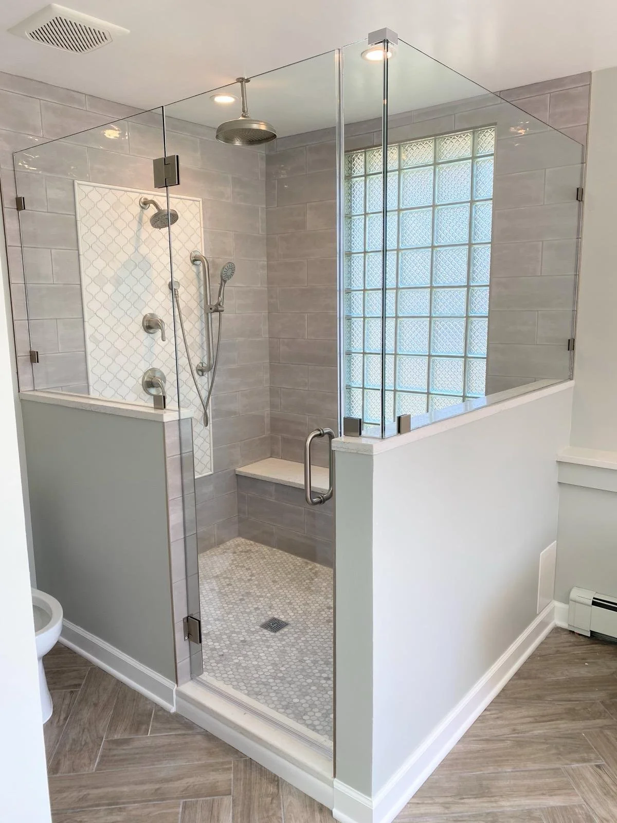Bathroom shower with glass enclosure, gray tiled walls, and a window with frosted glass block. The shower has a rain showerhead, a handheld shower, and multiple body jets. The floor is tiled with small hexagonal tiles, and there is a built-in bench i
