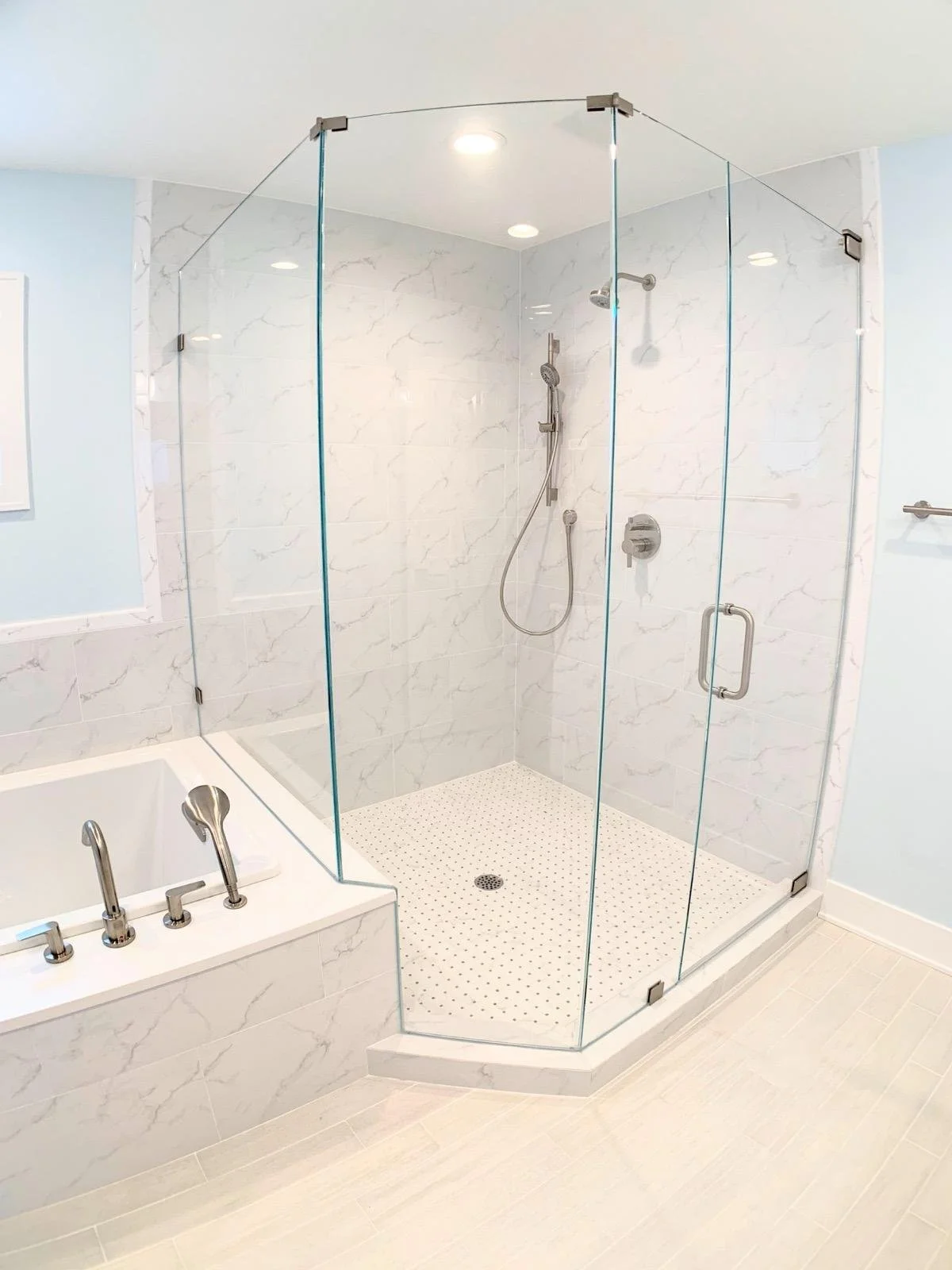 A modern bathroom features a glass shower enclosure with marble wall tiles, a handheld showerhead, and a built-in bathtub with chrome fixtures. Light-colored wood or tile flooring complements the aesthetic.