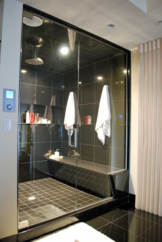 A modern glass-enclosed shower with black tiles, white towels hanging on hooks, and a built-in bench with toiletries.